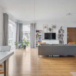 Adrian Tokarz Marketing - Jasne wnętrze salonu z drewnianą podłogą, szarą sofą i regałami z książkami. Widok na okno z szarymi zasłonami i roślinę doniczkową. Minimalistyczny stół z krzesłami.