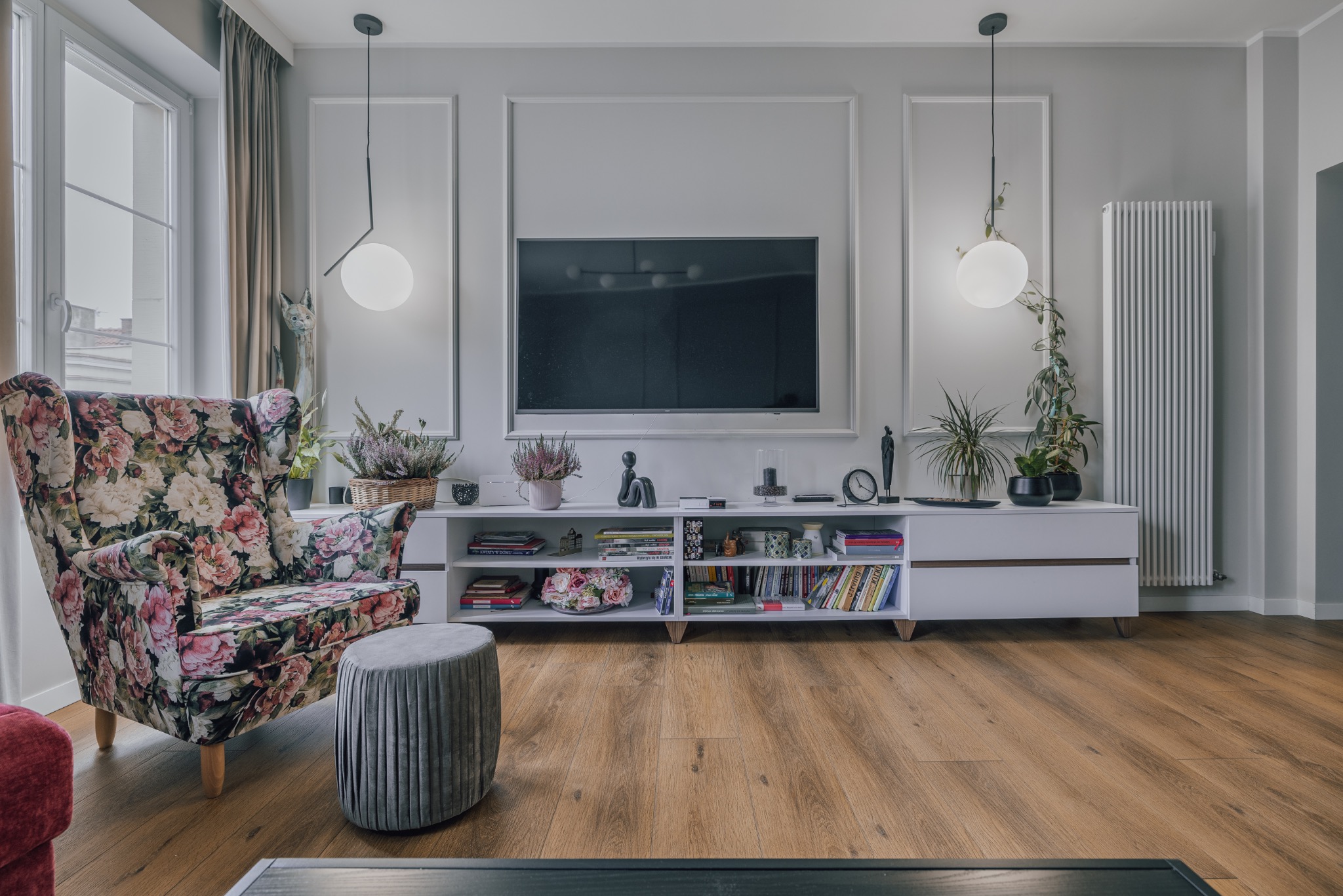 Salon w odcieniach szarości z drewnianą podłogą. W centrum biała szafka RTV z dekoracjami, nad nią telewizor. Po lewej fotel w kwiaty i szary puf.