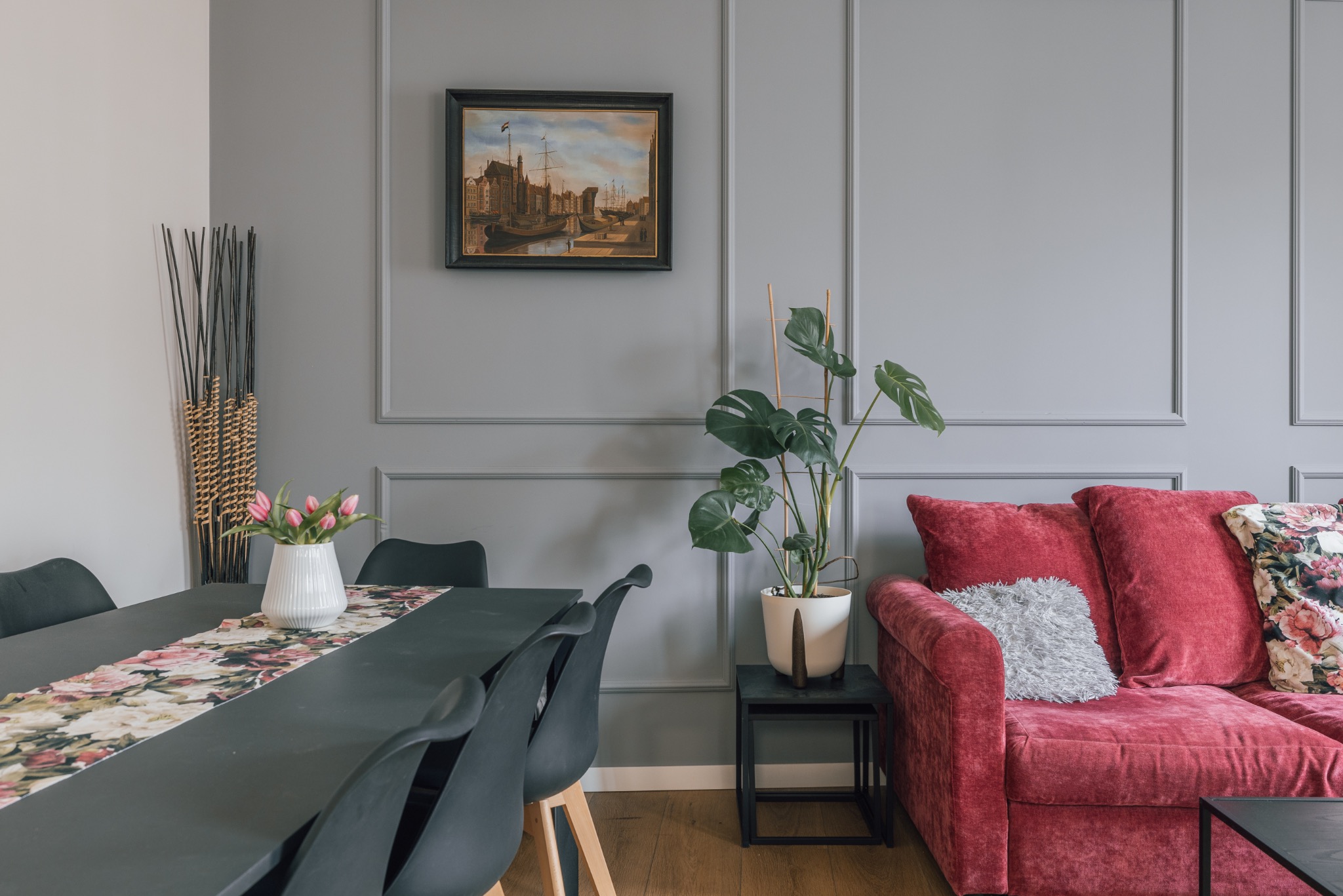 Stylowe wnętrze: szare ściany z dekoracyjnymi listwami, obraz z motywem morskim, czerwona sofa i czarny stół z kwiatami. Aranżacja z roślinami doniczkowymi.