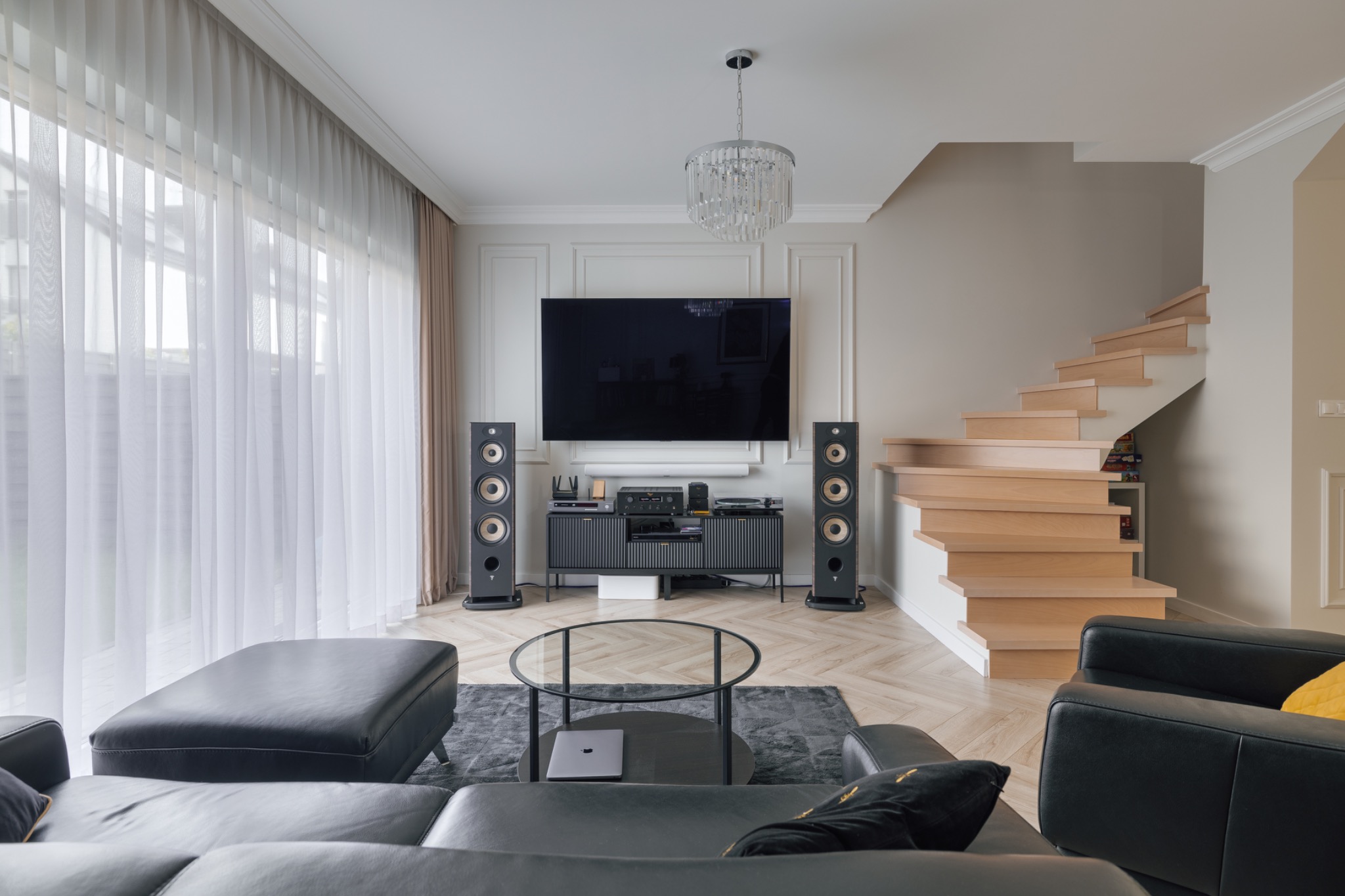 Elegancki salon z czarną skórzaną sofą, szklanym stolikiem kawowym i nowoczesnym sprzętem audio-video. Jasne drewniane schody i subtelne oświetlenie dopełniają wnętrze.