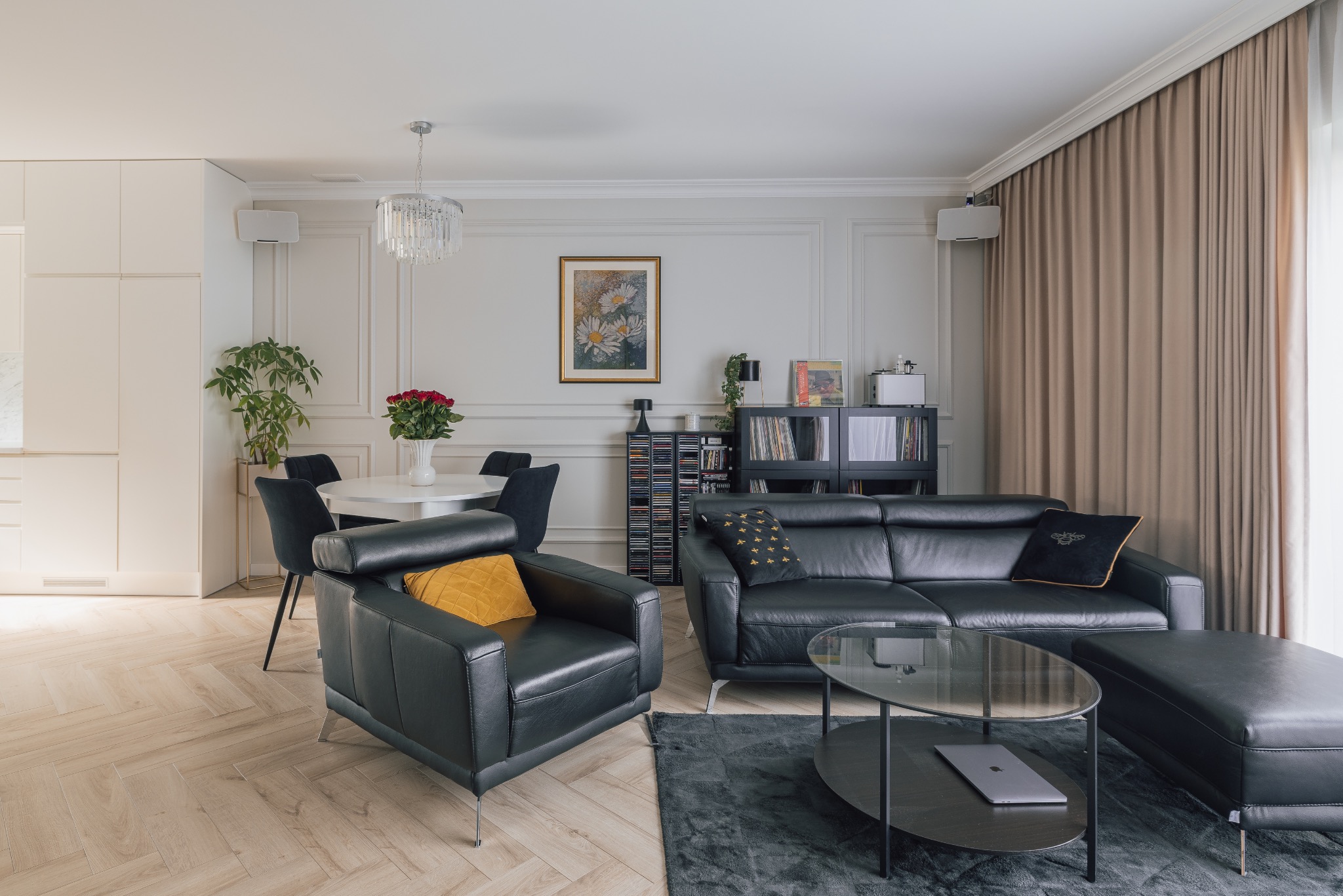 Elegancki salon z czarną skórzaną sofą, fotelem i szklanym stolikiem kawowym na tle jasnych ścian z dekoracyjnymi listwami i parkietu w jodełkę. W tle stół z kwiatami.