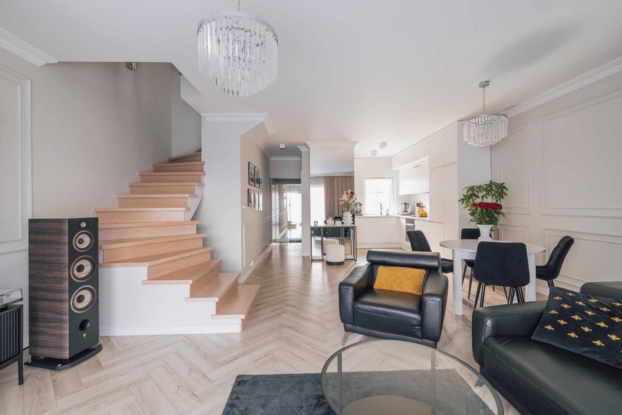 Jasne wnętrze domu z drewnianymi schodami, parkietem w jodełkę i czarnymi skórzanymi fotelami. Widoczna kuchnia i jadalnia w tle. Kryształowy żyrandol dodaje elegancji.