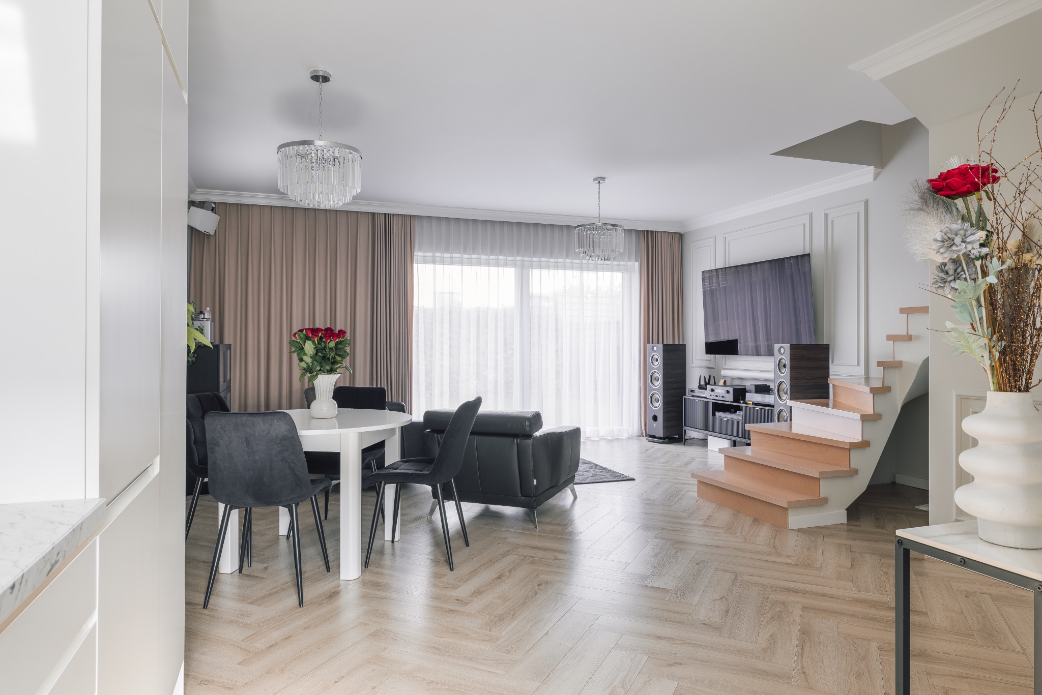 Jasne wnętrze salonu z parkietem w jodełkę, okrągłym stołem i czarnymi krzesłami. W tle schody, telewizor i kolumny głośnikowe. Dwa kryształowe żyrandole.