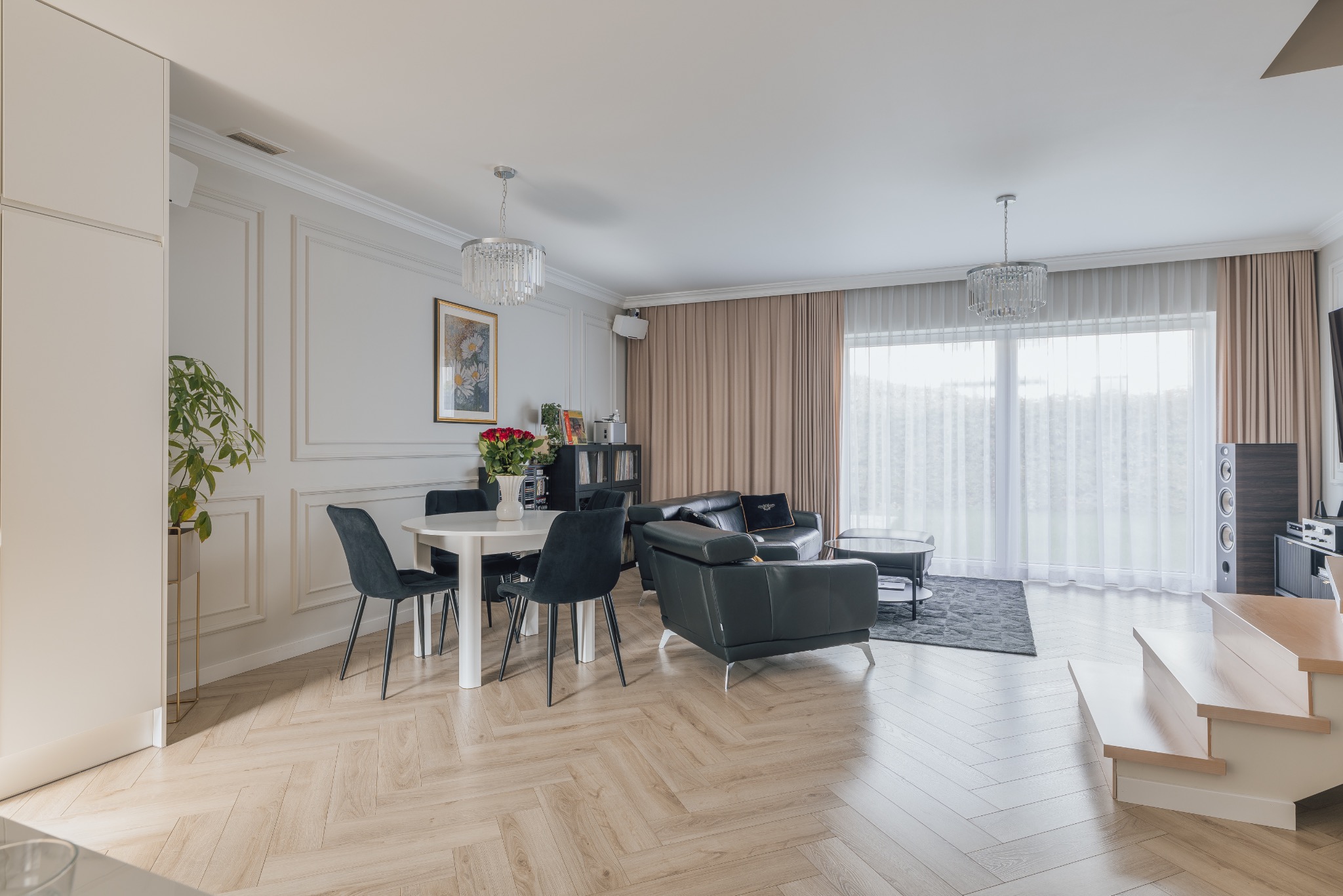 Jasne wnętrze salonu z parkietem w jodełkę, okrągłym stołem i czarnymi krzesłami. W tle skórzana sofa, okno z zasłonami i schody. Całość w neutralnej kolorystyce.
