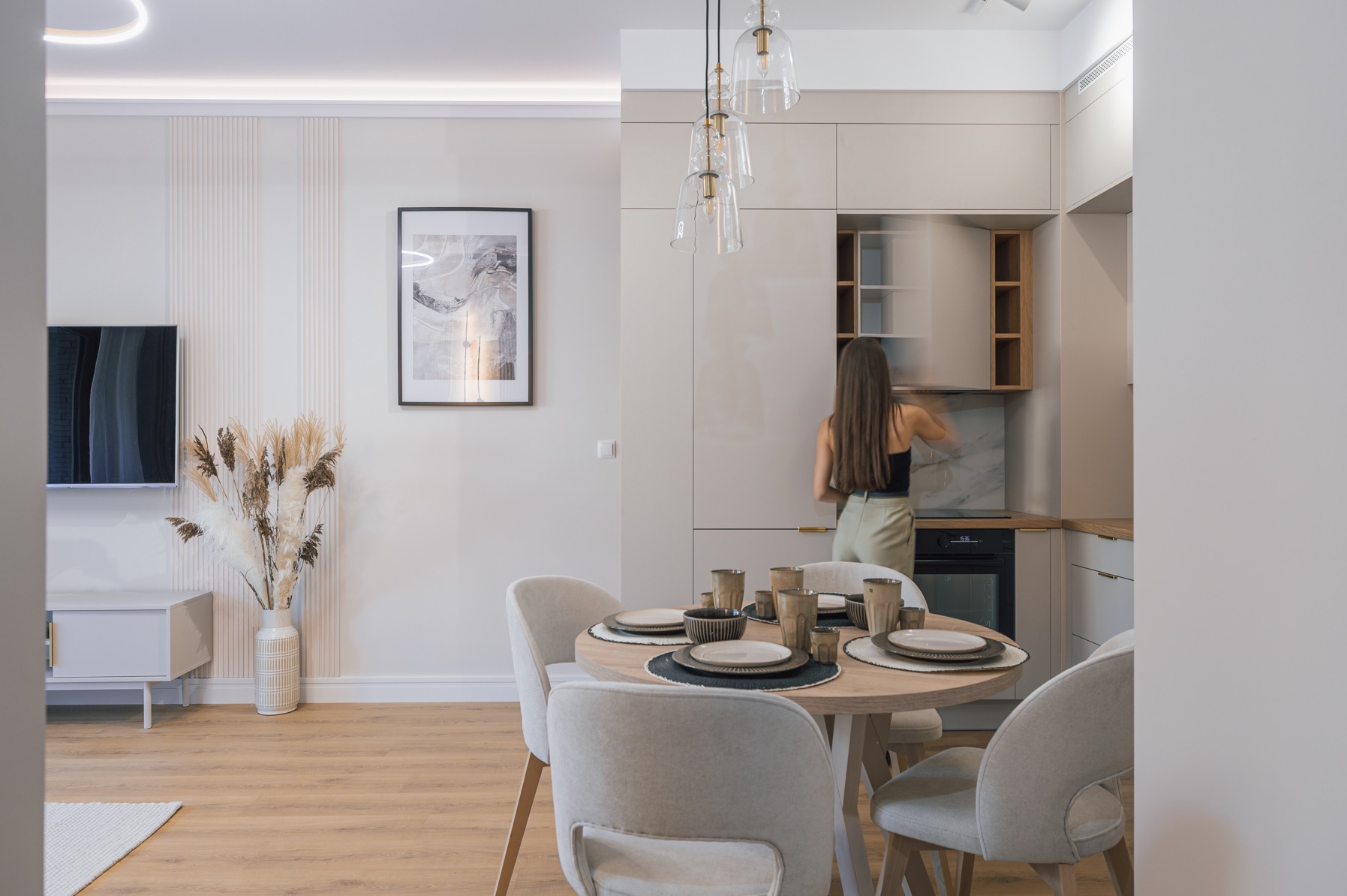 Jasne wnętrze: okrągły stół z nakryciem, krzesła, szafki kuchenne z otwartą szafką, kobieta sięga do szafki, dekoracje z suszonych traw, telewizor na ścianie.