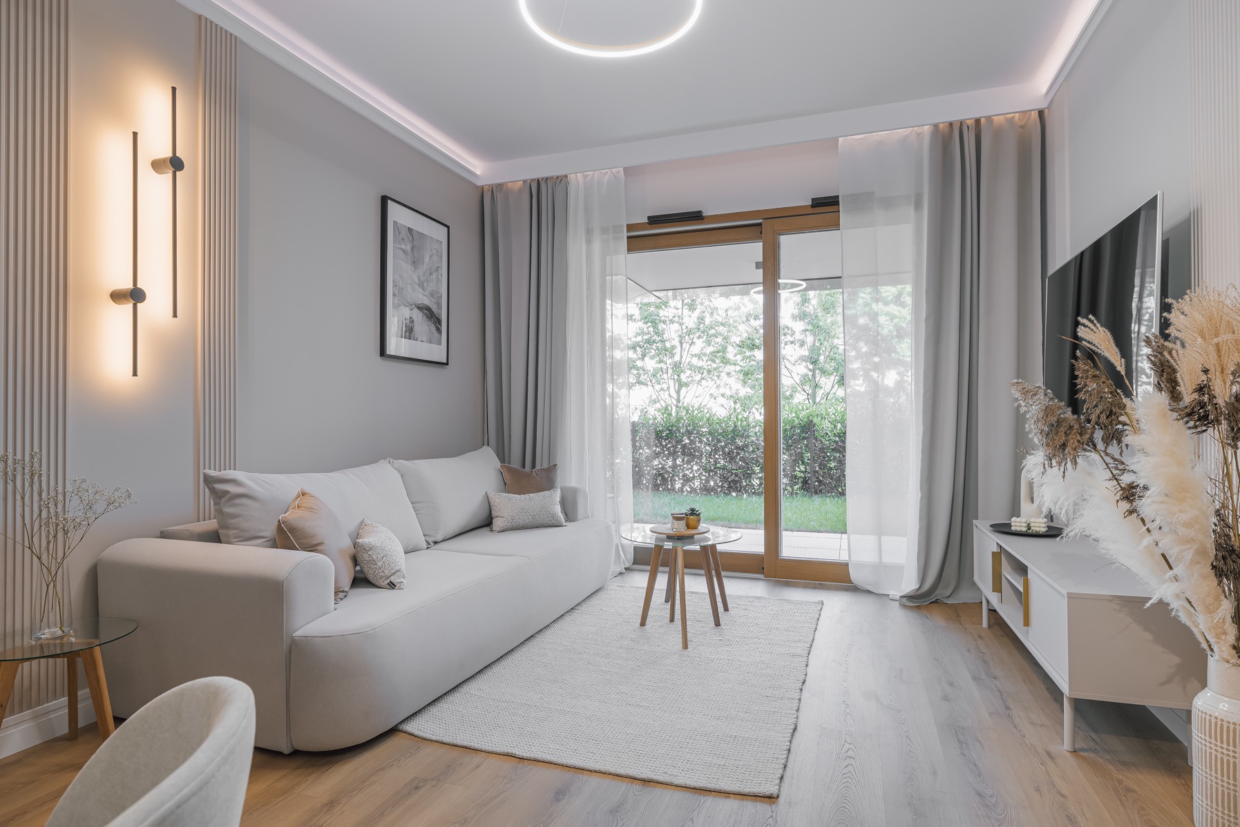 Jasny salon z beżową sofą, drewnianą podłogą i dużym oknem z widokiem na ogród. Minimalistyczny design z okrągłą lampą sufitową i dekoracyjnymi trawami w wazonie.