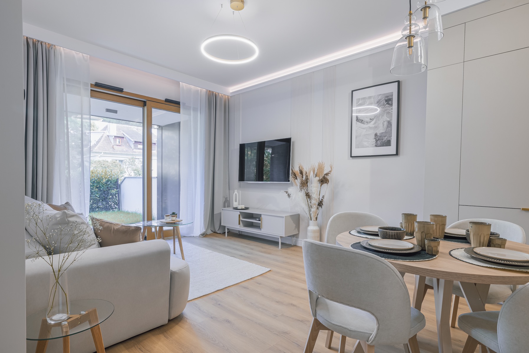 Jasne wnętrze: salon z jadalnią w neutralnych barwach. Sofa, okrągły stół z nakryciem, telewizor na tle ściany z dekoracyjnymi panelami. Duże okno z widokiem na zieleń.