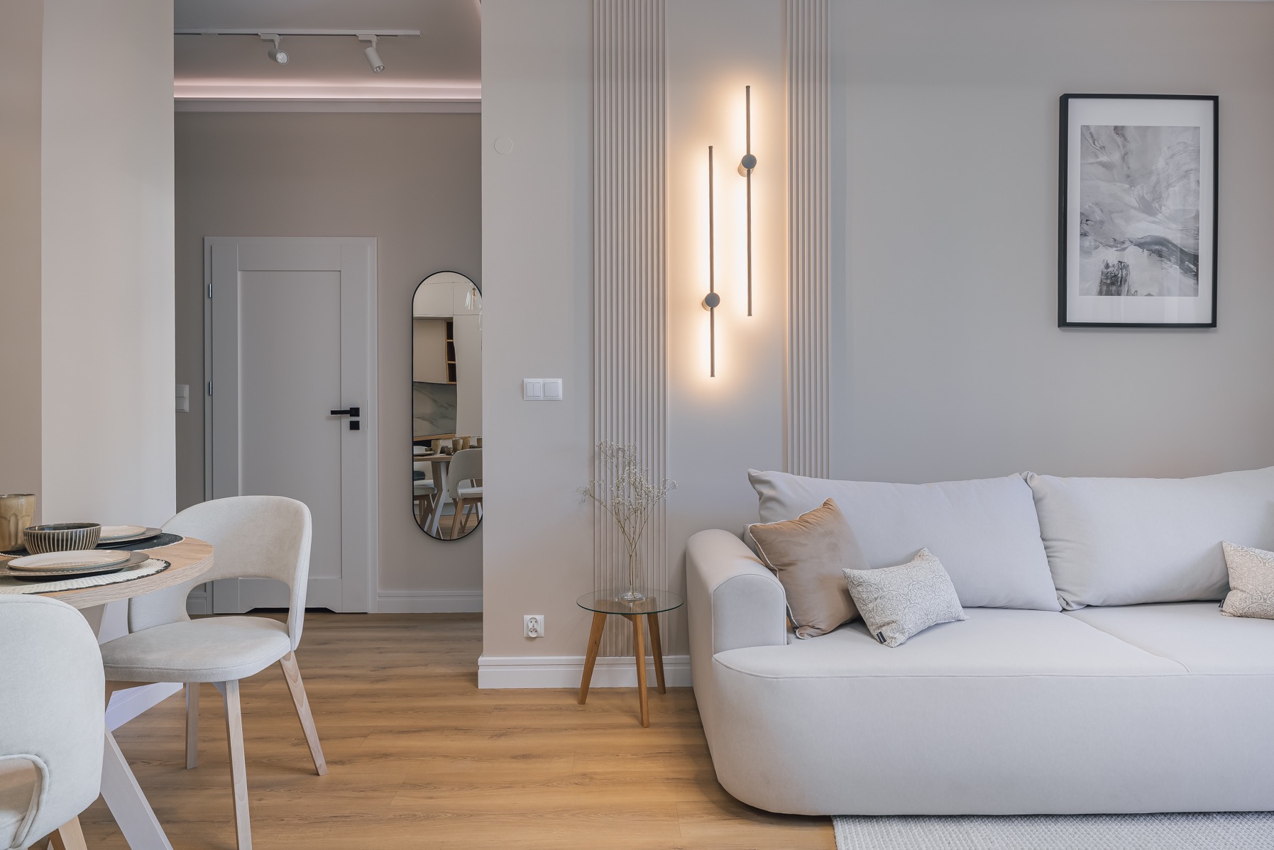 Jasne wnętrze: sofa, stół z krzesłami, minimalistyczna lampa ścienna i lustro owalne. Neutralna kolorystyka, drewniana podłoga, nowoczesny design.