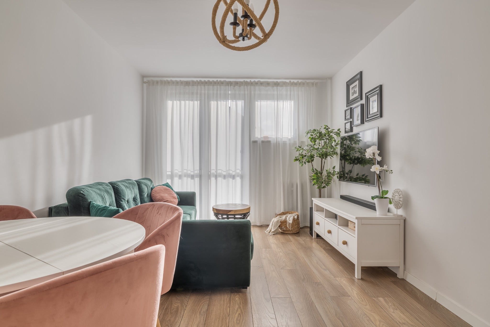 Jasne wnętrze salonu z zieloną sofą, różowymi krzesłami i drewnianą podłogą. Dekoracje obejmują lampę z liny, rośliny i galerię zdjęć na ścianie.