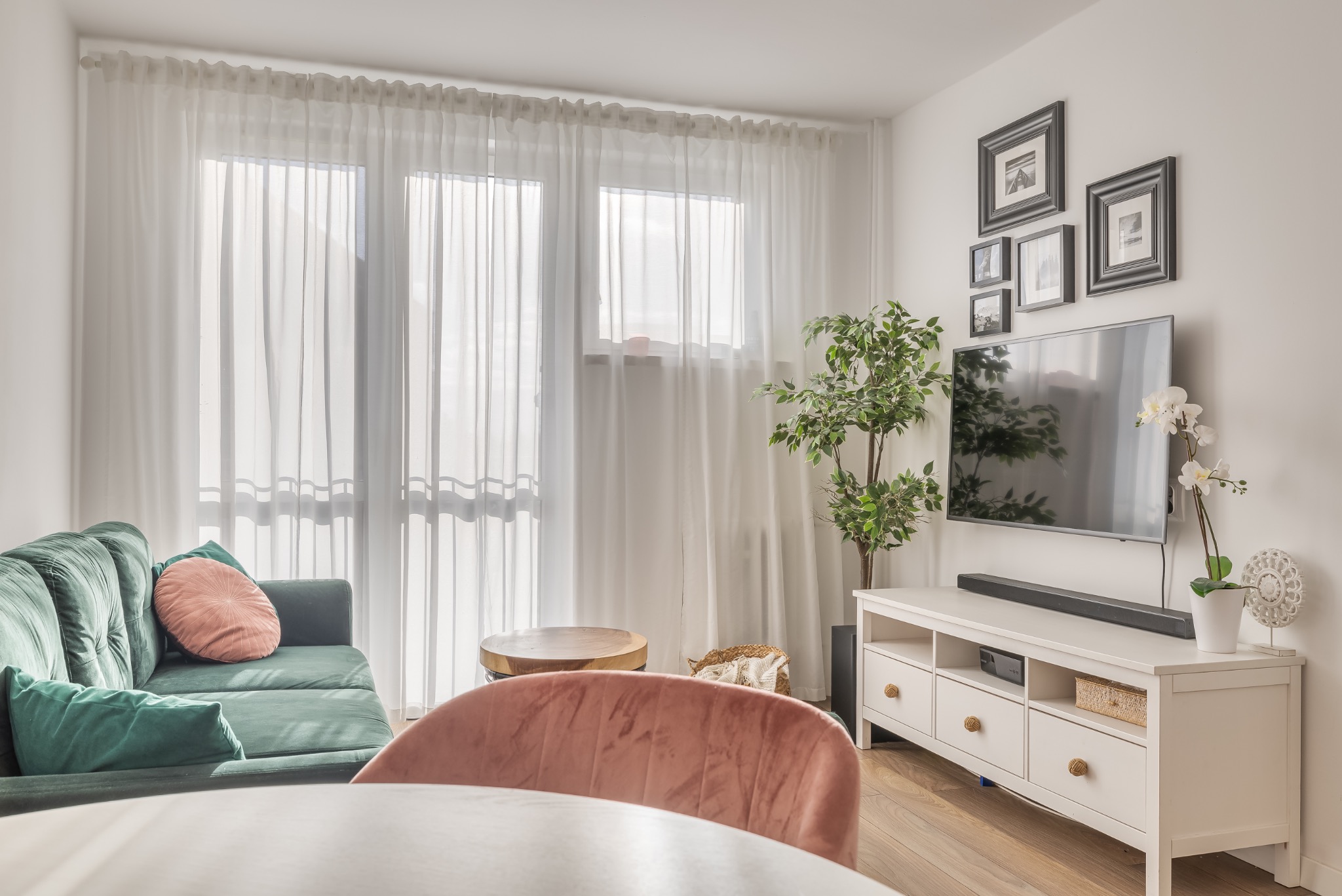 Jasne wnętrze salonu z zieloną sofą, telewizorem i dekoracjami. Widoczne okno z białymi zasłonami i drewniany stolik kawowy. Ustawienie mebli i dodatków tworzy przytulną atmosferę.