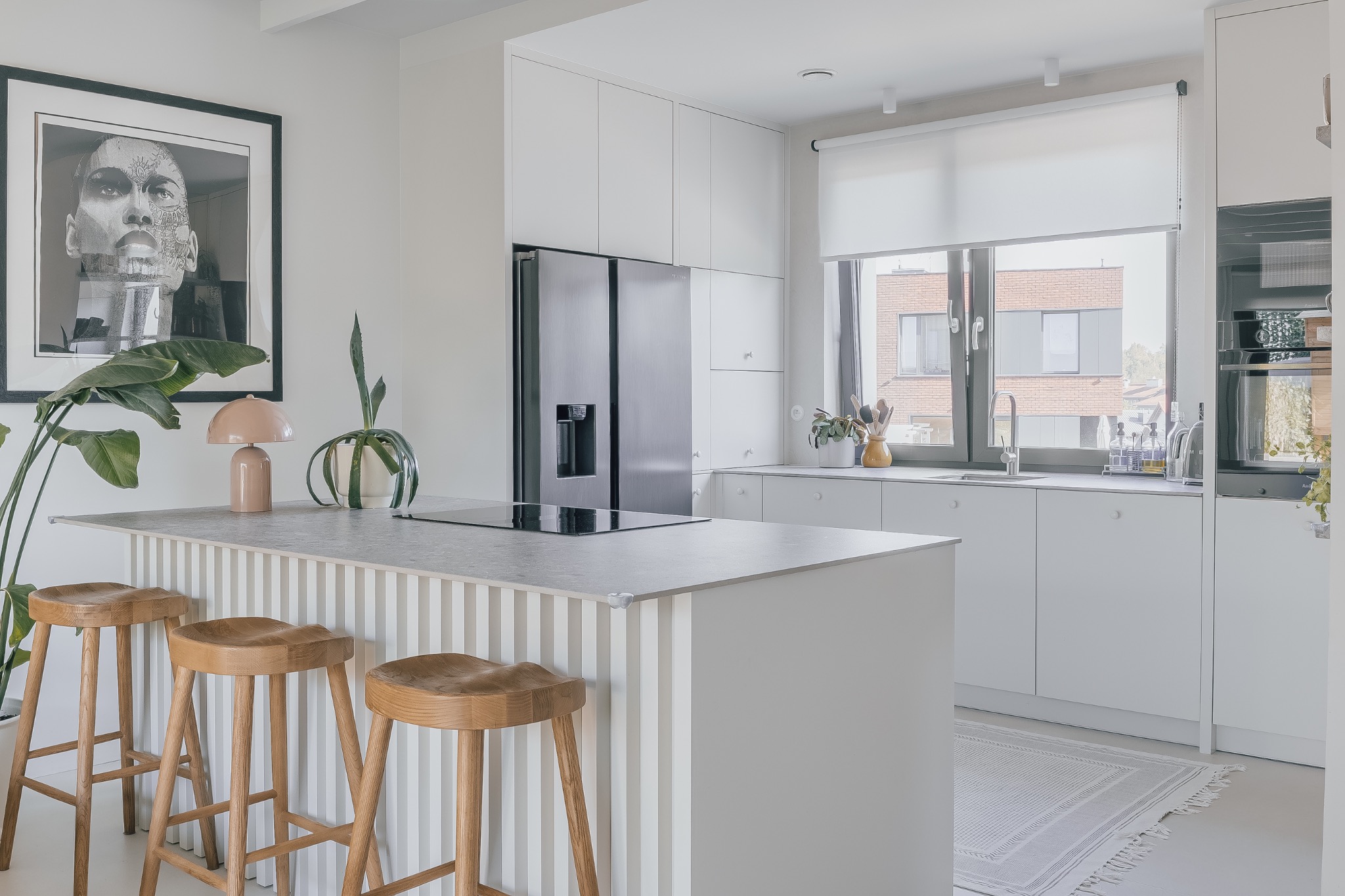 Jasna kuchnia w minimalistycznym stylu: białe szafki, wyspa z hokerami, czarna lodówka side-by-side i grafika na ścianie. Nowoczesne oświetlenie i rośliny.