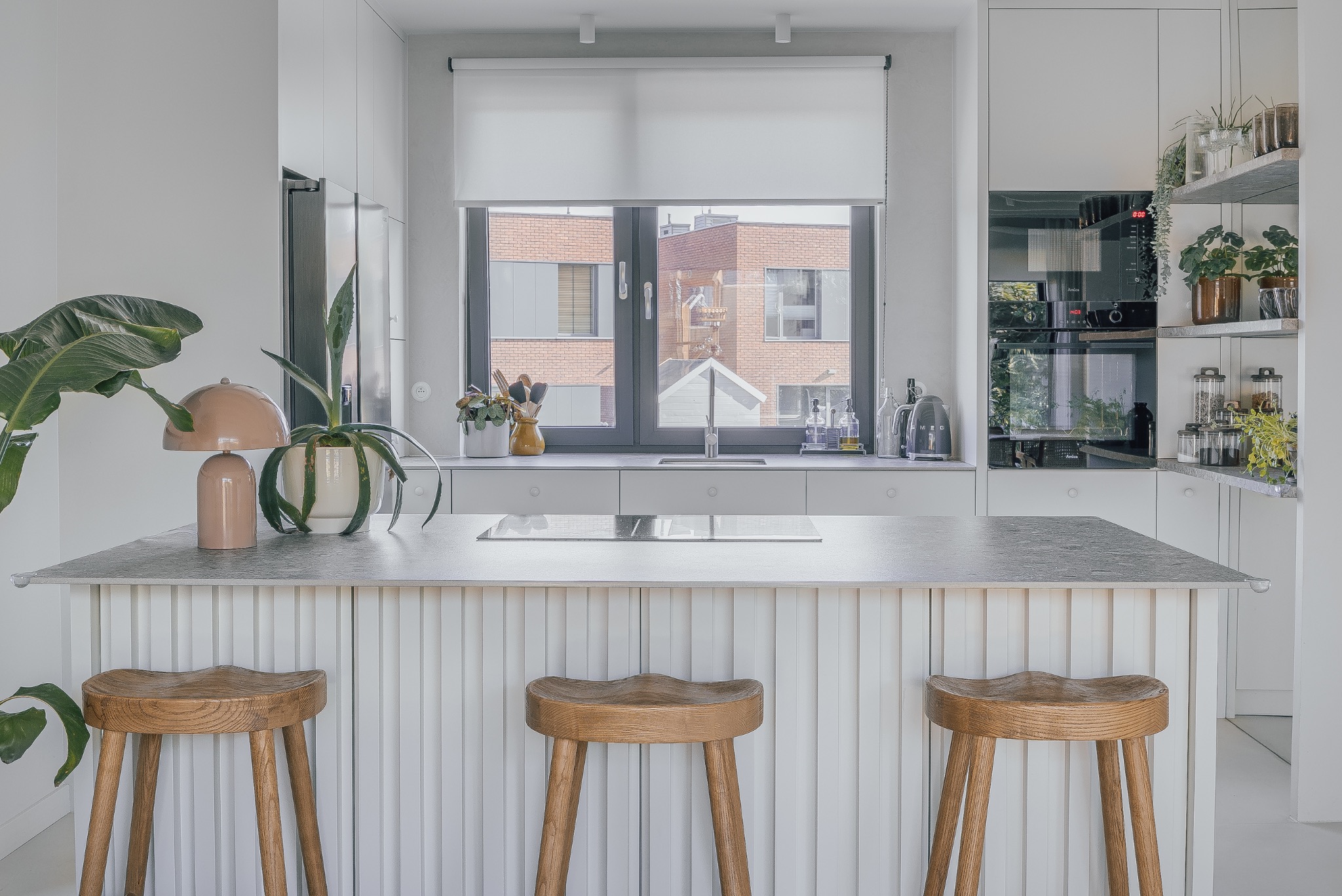 Jasna kuchnia w stylu skandynawskim z wyspą, drewnianymi stołkami barowymi i roślinami doniczkowymi. Widok na budynek z cegły za oknem.