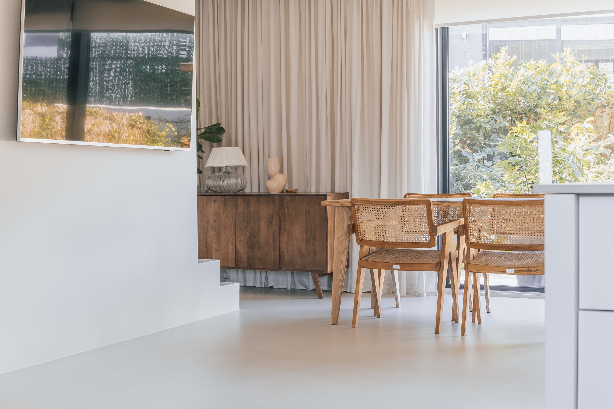 Jasne wnętrze z drewnianym kredensem, lampą i krzesłami z rattanu. Za oknem zieleń, a na ścianie telewizor. Minimalistyczny design z naturalnymi akcentami.