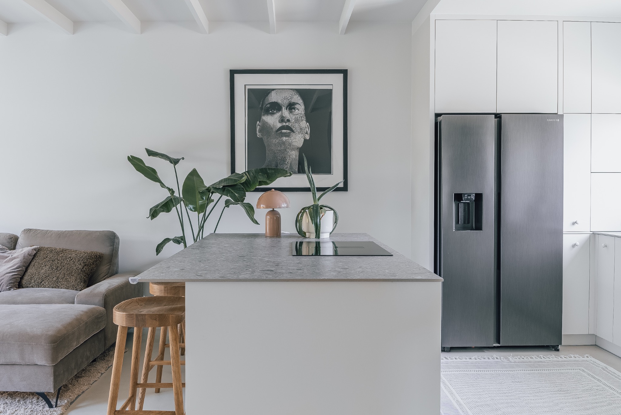 Nowoczesne wnętrze: szara sofa, drewniane stołki barowe, wyspa kuchenna z blatem imitującym kamień, czarno-biały portret na ścianie i srebrna lodówka side-by-side.
