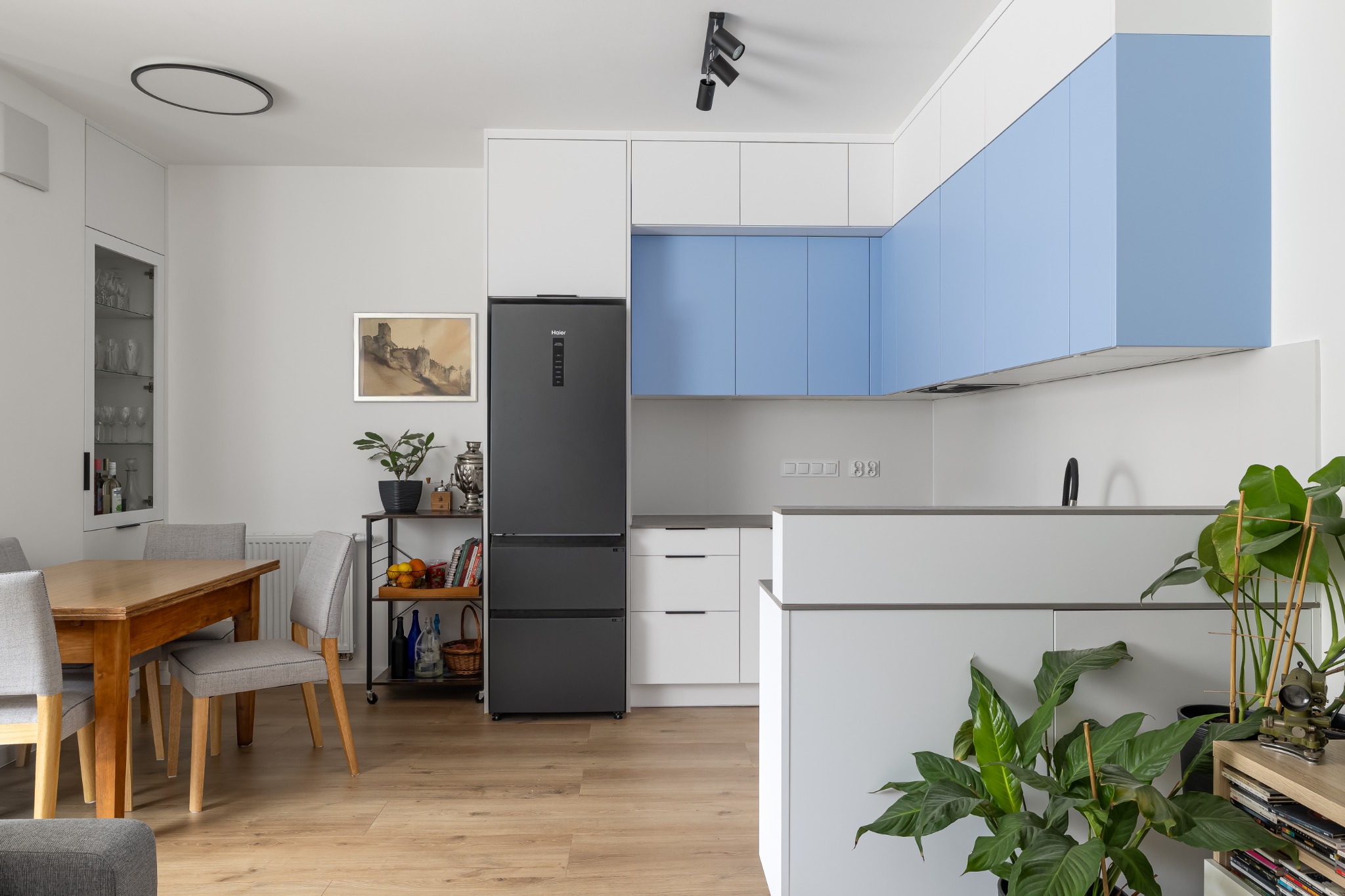 Jasne wnętrze kuchni z drewnianym stołem i szarymi krzesłami. W tle zabudowa kuchenna z błękitnymi szafkami i czarna lodówka. Na podłodze panele imitujące drewno.