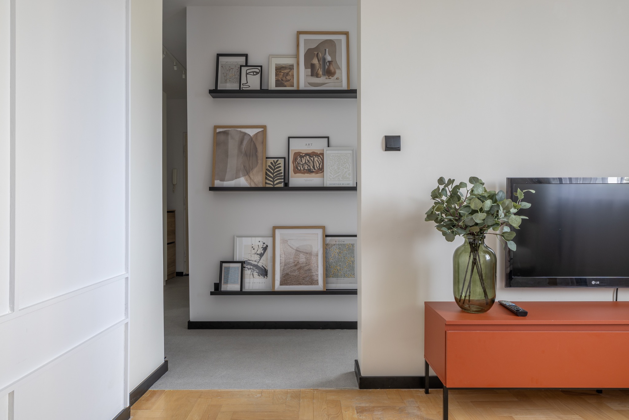 Jasne wnętrze: minimalistyczny salon z drewnianą podłogą, półkami z grafikami, pomarańczową komodą i telewizorem. Wazon z zielonymi gałązkami dodaje naturalnego akcentu.