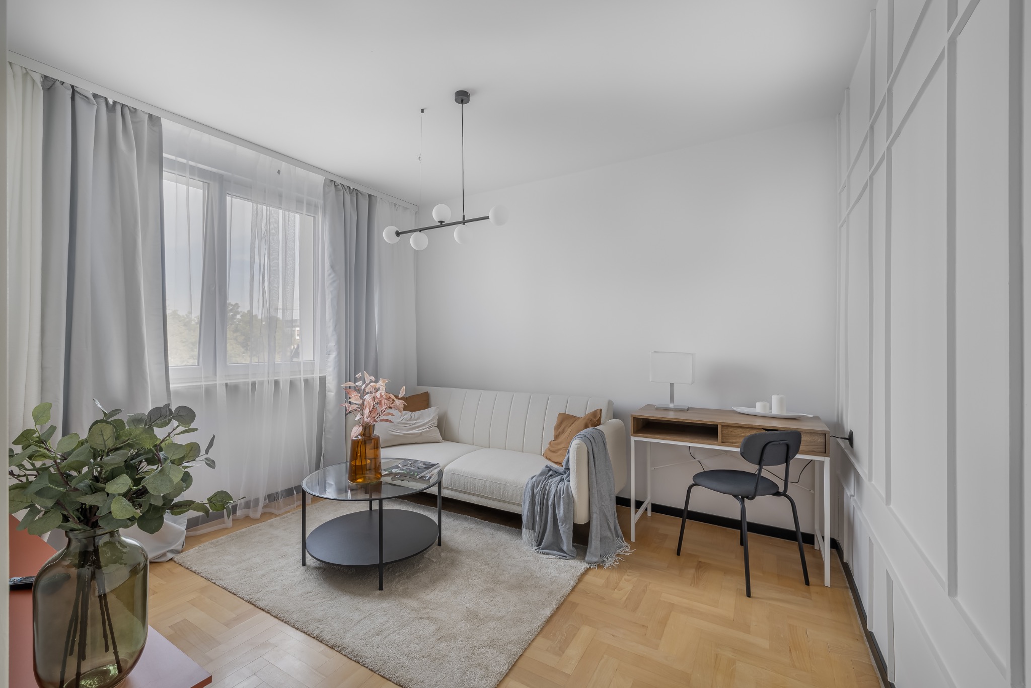 Jasne wnętrze salonu z parkietem w jodełkę, sofą, stolikiem kawowym i biurkiem. Stonowana kolorystyka, minimalistyczny design, dekoracyjne panele ścienne.