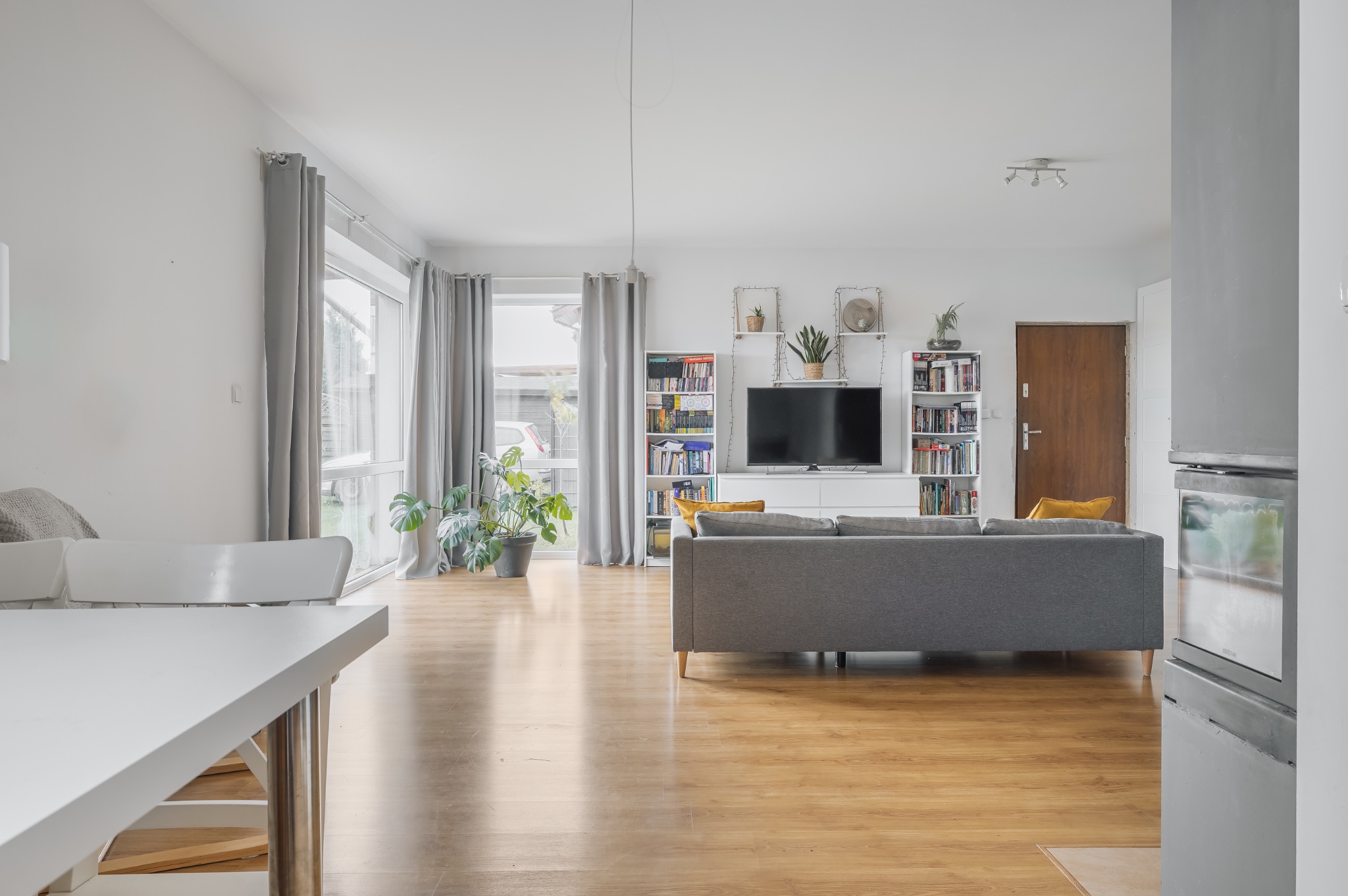 Jasne wnętrze salonu z drewnianą podłogą, szarą sofą i regałami z książkami. Widok na okno z szarymi zasłonami i roślinę doniczkową. Minimalistyczny stół z krzesłami.