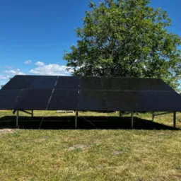 Czarny panel fotowoltaiczny na metalowej konstrukcji, ustawiony na trawiastym terenie z drzewem w tle, pod błękitnym niebem z chmurami.