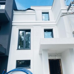 PORTBUD Kyrylo Portnov - Biała elewacja budynku z ciemnymi oknami w nowoczesnym stylu. Widoczne rusztowanie i niebieskie rury. Częściowo zachmurzone niebo.