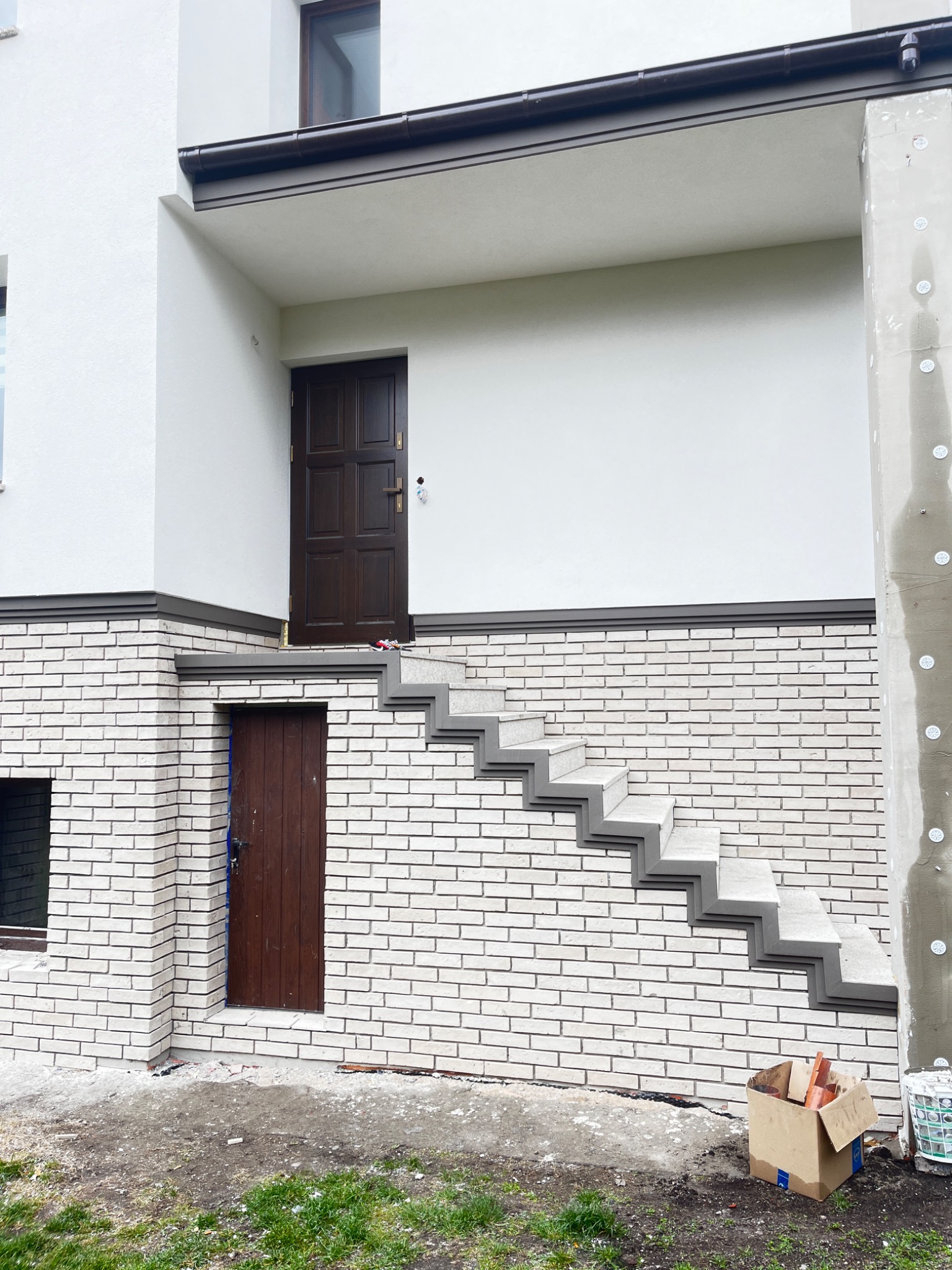 Biała ceglana elewacja z nietypowymi schodami zewnętrznymi prowadzącymi do brązowych drzwi. Fragment ocieplonego słupa z boku. Karton z resztkami materiałów budowlanych.