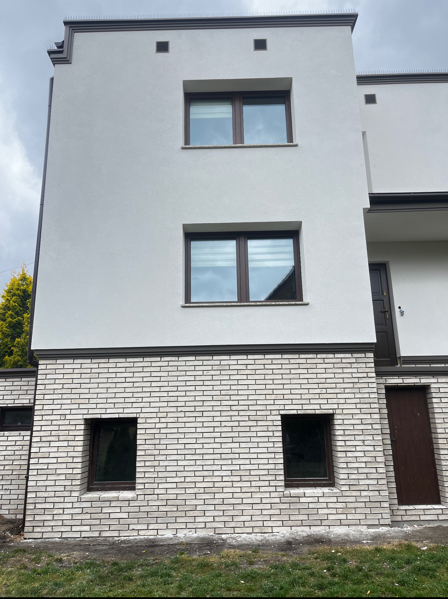 Nowoczesna elewacja budynku z białym tynkiem i ceglanym cokołem. Widoczne okna z brązowymi ramami i zabezpieczenia przeciw ptakom na dachu. Ujęcie z dołu.