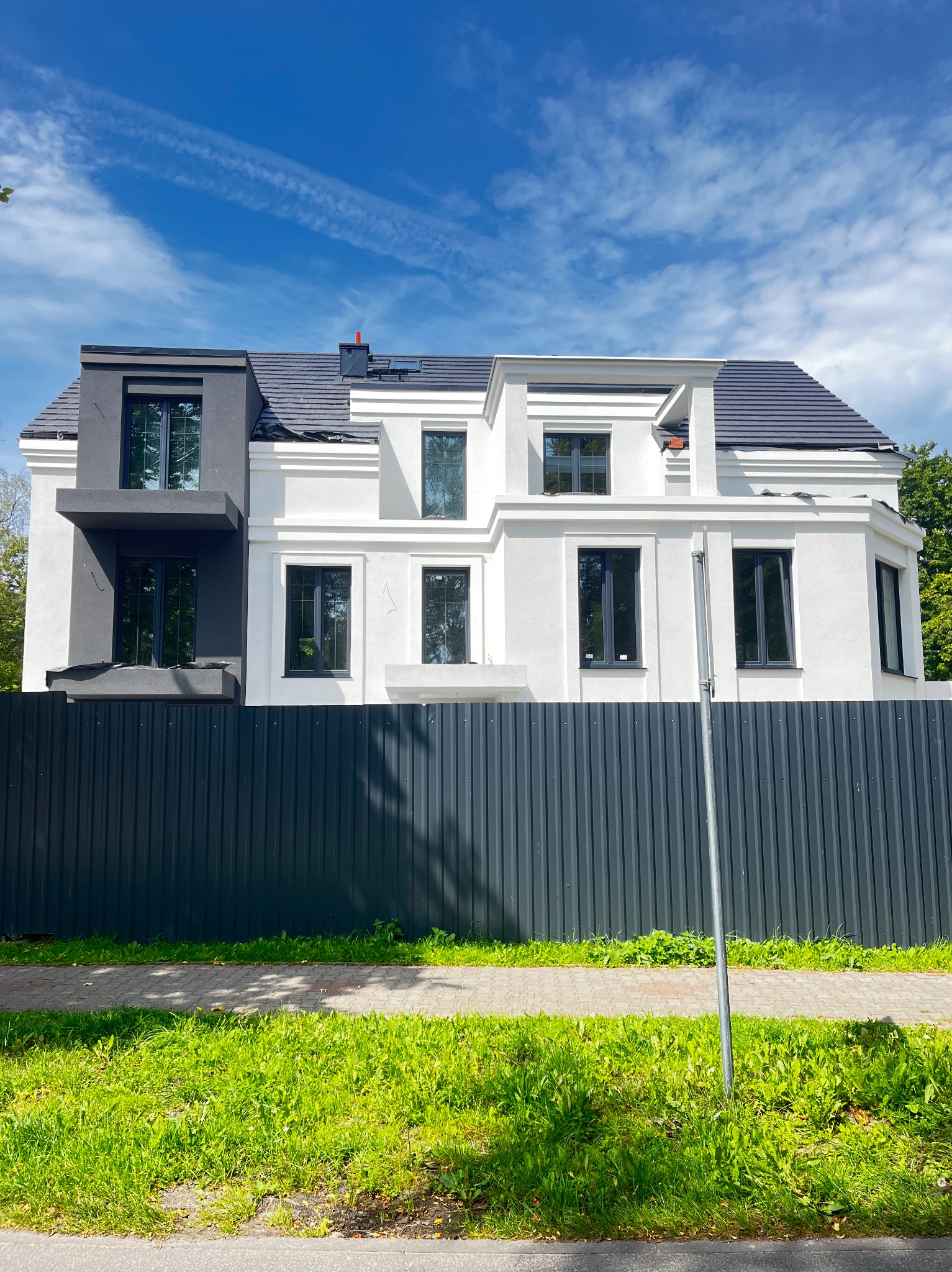 Dwukolorowy dom z szarą elewacją po lewej i białą po prawej stronie, widoczny zza ciemnoszarego, metalowego ogrodzenia. Błękitne niebo z chmurami w tle.