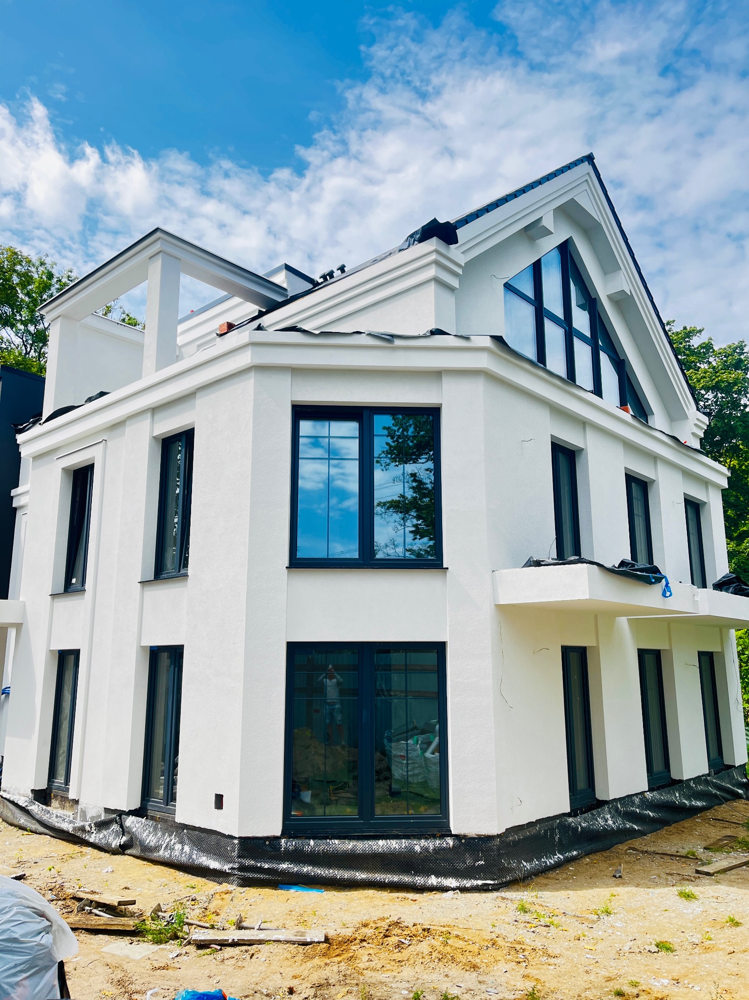 Elegancki, dwukondygnacyjny dom z białym, świeżo położonym tynkiem, ciemnymi oknami i fragmentem czarnej folii hydroizolacyjnej u podstawy elewacji.