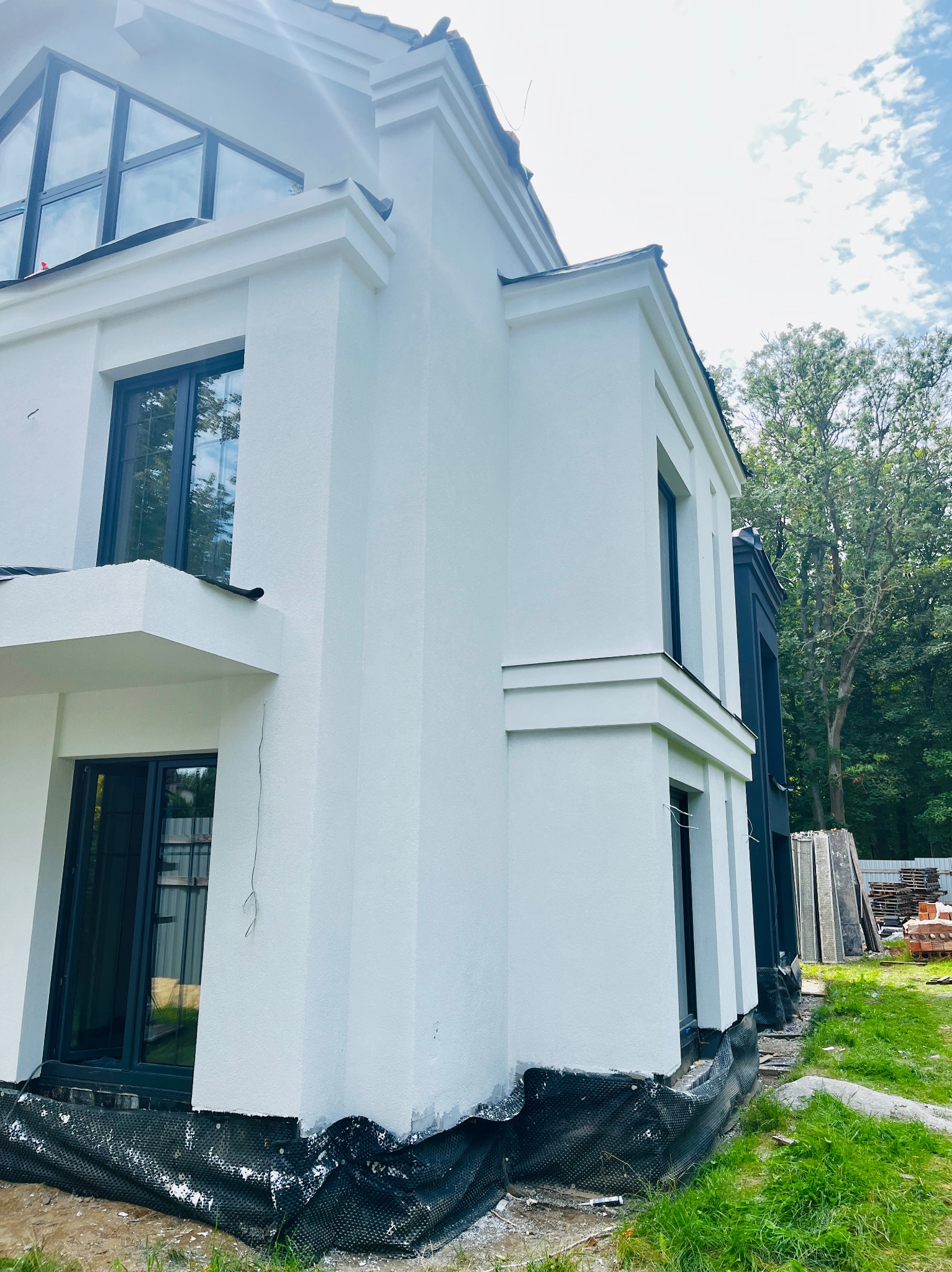 Nowoczesny dom z białą elewacją, czarnymi oknami i dekoracyjnymi elementami architektonicznymi. Widoczny fragment ogrodu i materiały budowlane.