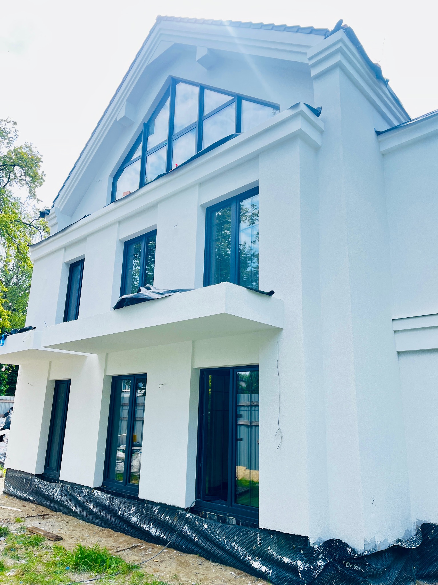 Nowoczesny, biały budynek z dużymi oknami i wykończoną elewacją. Widoczne zabezpieczenia przy gruncie, sugerujące prace wykończeniowe.