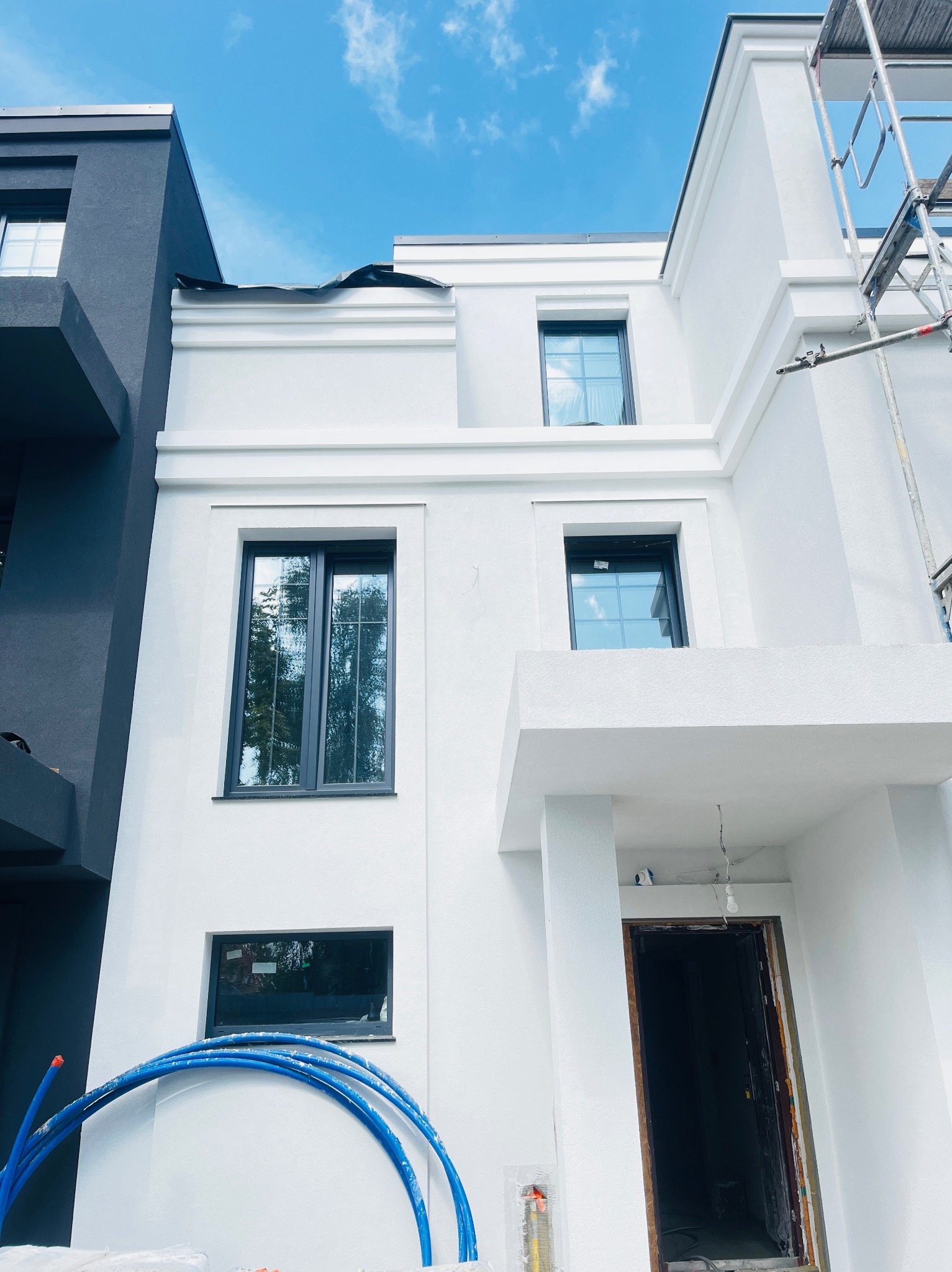 Biała elewacja budynku z ciemnymi oknami w nowoczesnym stylu. Widoczne rusztowanie i niebieskie rury. Częściowo zachmurzone niebo.