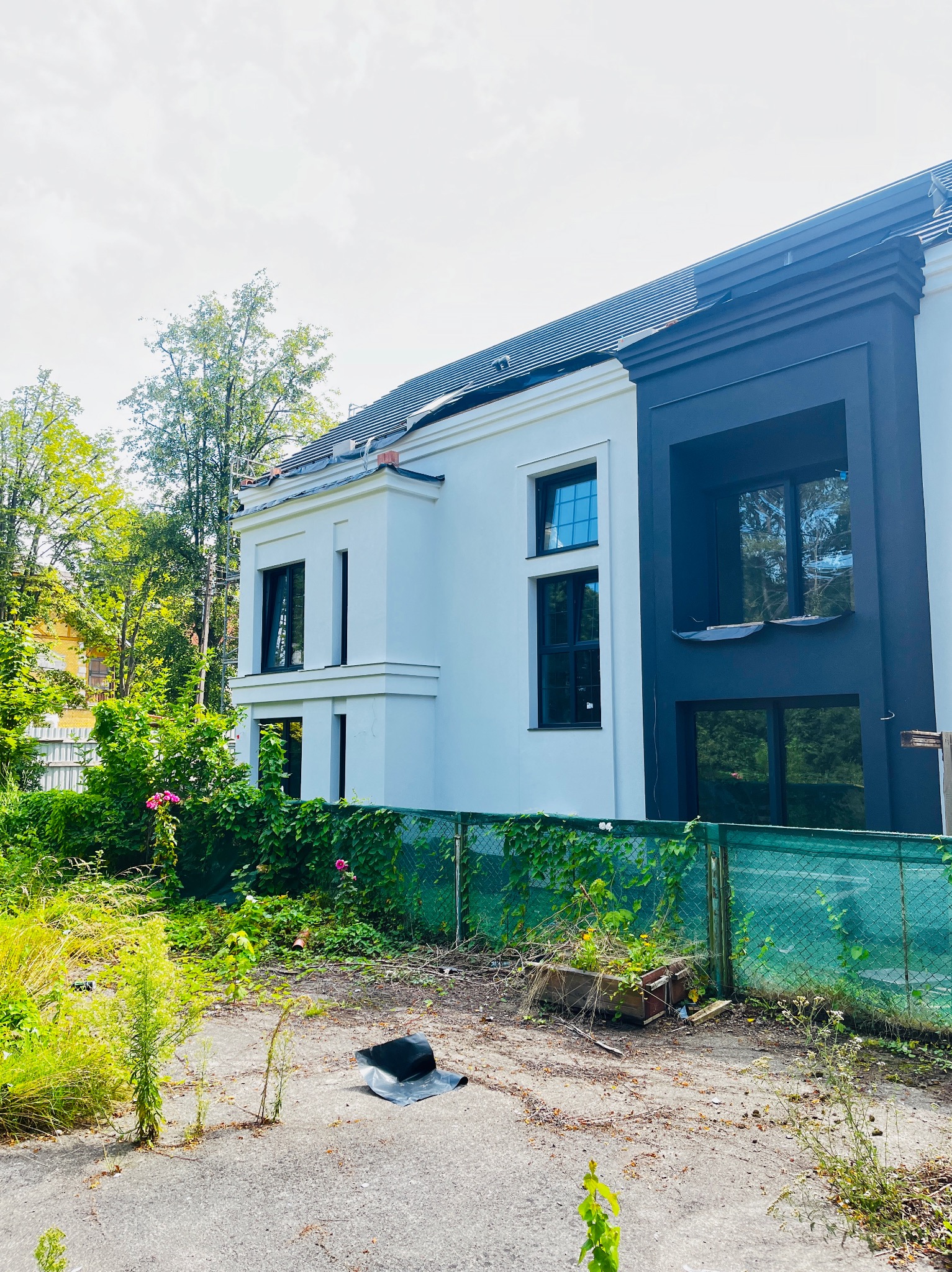 Nowoczesny dom z biało-czarną elewacją, widoczny przez zielone ogrodzenie. Część dachu w trakcie prac. Zarośnięty teren przed budynkiem. Słoneczny dzień.