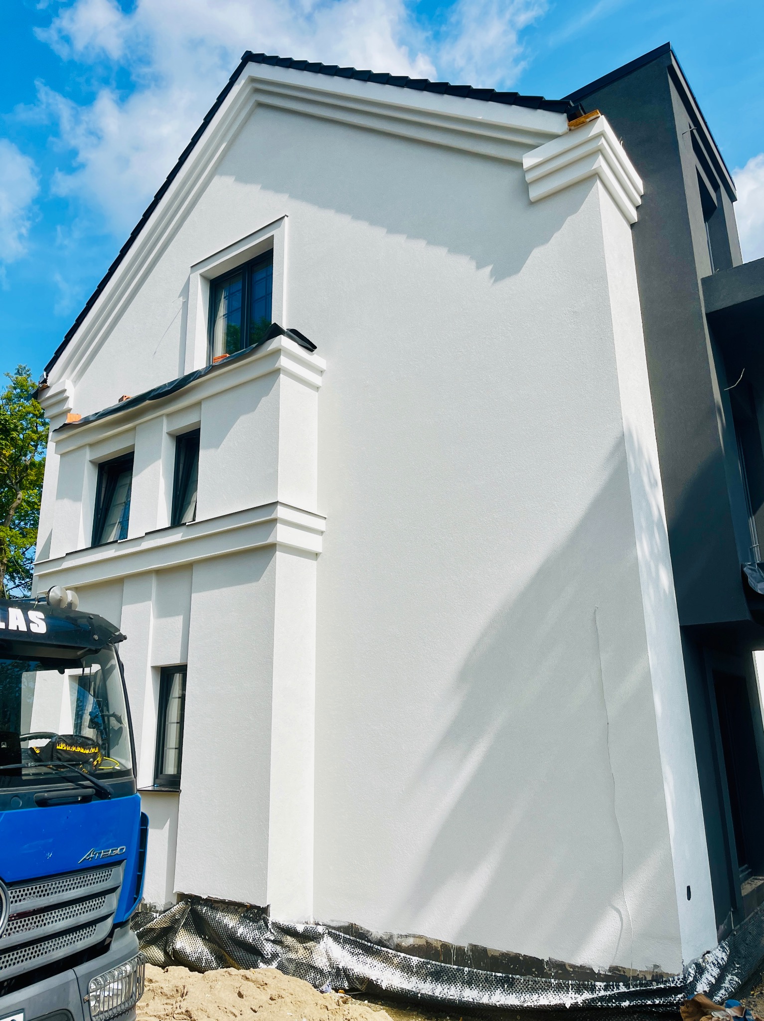 Biała elewacja domu jednorodzinnego z niebieską ciężarówką w kadrze. Widoczne detale architektoniczne i tło z błękitnym niebem. Nowoczesny design.