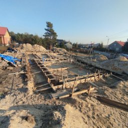 S bud dach - Szeroki kadr na wykop pod fundamenty domu jednorodzinnego z drewnianym szalunkiem, z kupami ziemi po bokach wykopu i okoliczną zabudową w tle.