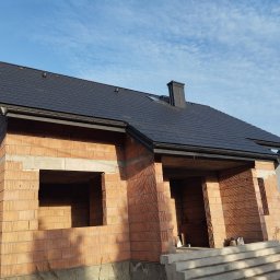 S bud dach - Surowy stan domu z czerwonej cegły ceramicznej z czarnym dachem na tle błękitnego nieba. Widoczne otwory okienne i drzwiowe, schody wejściowe.