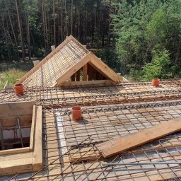 S bud dach - Zbrojenie stropu z drewnianą konstrukcją więźby dachowej na tle lasu, widok z góry. Prace budowlane w toku, zbrojenie z siatki stalowej, drewniane szalunki.