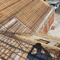 S bud dach - Zbrojenie stropu na drewnianym szalunku, widok z góry. Na drugim planie ściana z czerwonej cegły i biały samochód dostawczy. Cień osoby na pierwszym planie.