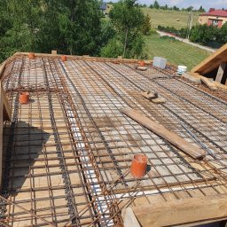 S bud dach - Zbrojenie dachu z siatki stalowej, drewniane szalunki, rury wentylacyjne. Widok z góry na konstrukcję przygotowaną do zalania betonem, w tle zieleń i zabudowa.