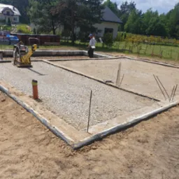 Przygotowany teren pod fundamenty domu jednorodzinnego. Ubita warstwa żwiru, widoczne szalunki i zbrojenia. W tle budynki mieszkalne i drzewa.