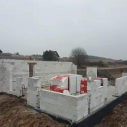Budowa domu z betonu komórkowego: ściany parteru wzniesione, palety z bloczkami Termobet na fundamencie. Widok na posesję i pochmurne niebo.
