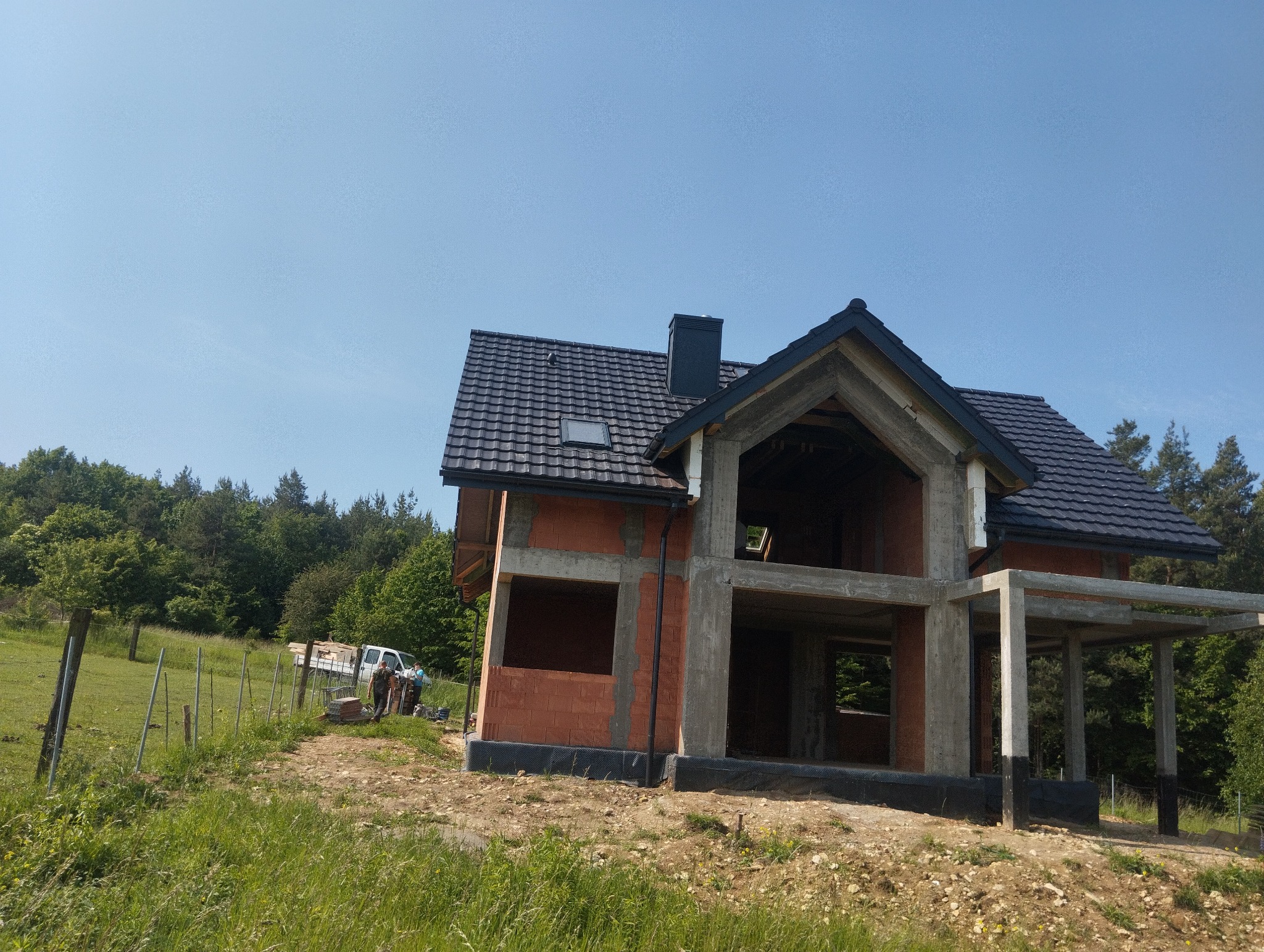 Budowa domu jednorodzinnego w stanie surowym otwartym, z widoczną konstrukcją ścian z pustaków i betonowych słupów, na tle zieleni i błękitnego nieba. Dach pokryty ciemną dachówką.