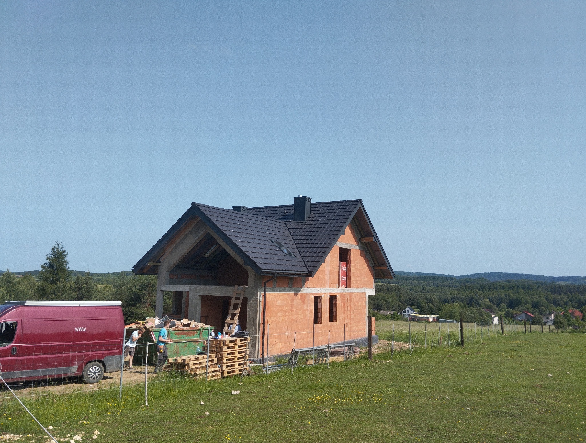 Dom w trakcie budowy z czerwonej cegły, z czarnym dachem i widocznymi pracownikami oraz kontenerem na odpady, na tle zielonej łąki i lasu.