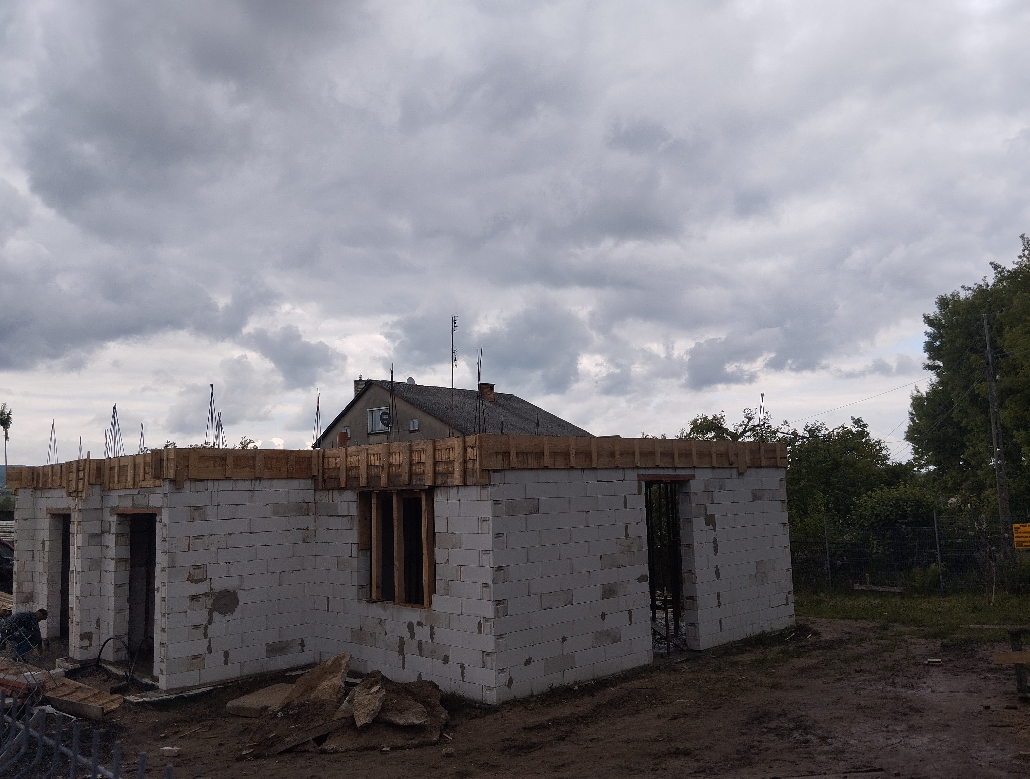 Wznoszenie ścian parterowego domu z betonu komórkowego. Widoczne drewniane szalunki. W tle pochmurne niebo i fragment starszego budynku.