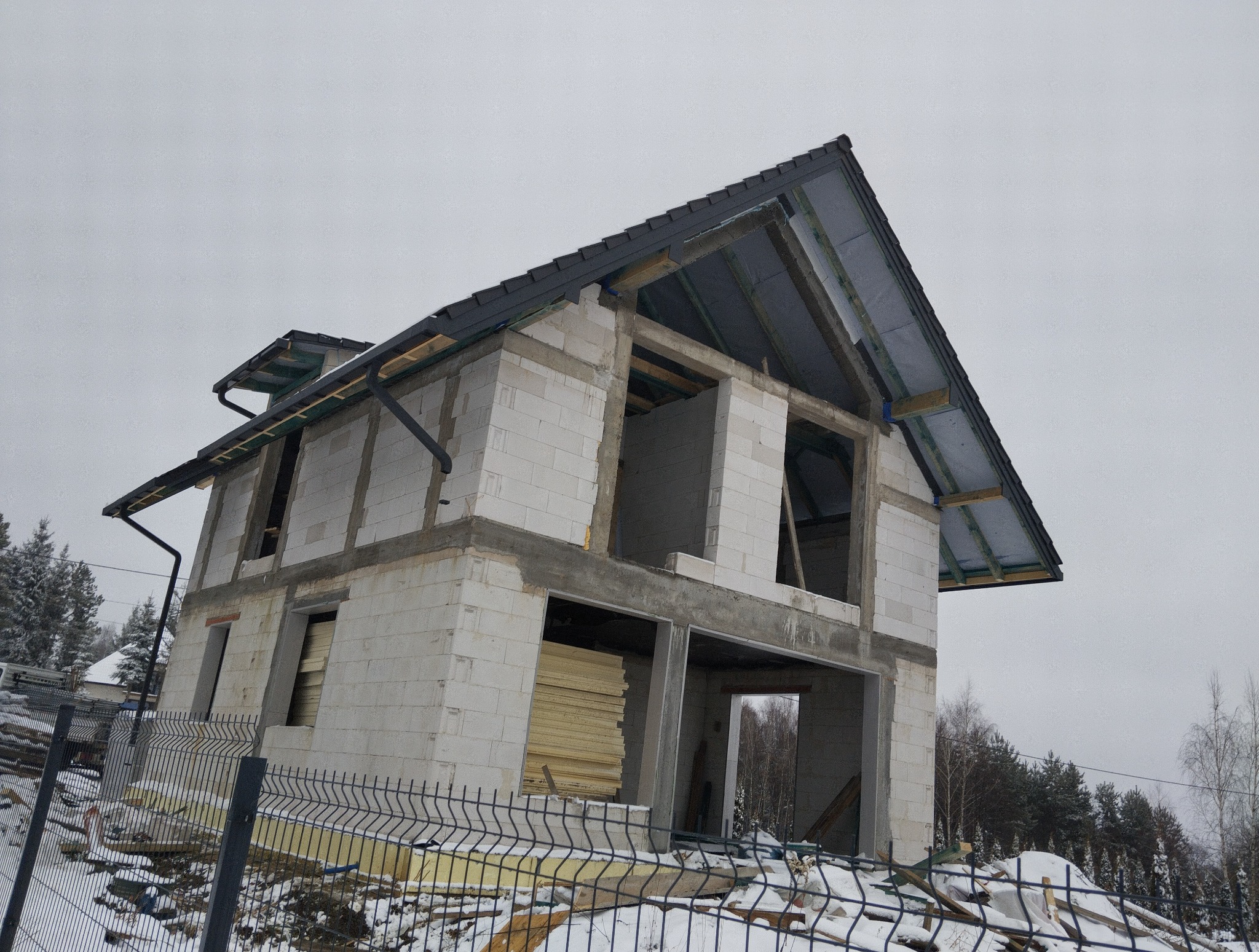 Surowy dom z białego pustaka w zimowym krajobrazie, widok pod kątem. Częściowo zadaszony, widoczne drewniane elementy konstrukcji dachu i okna bez szyb.