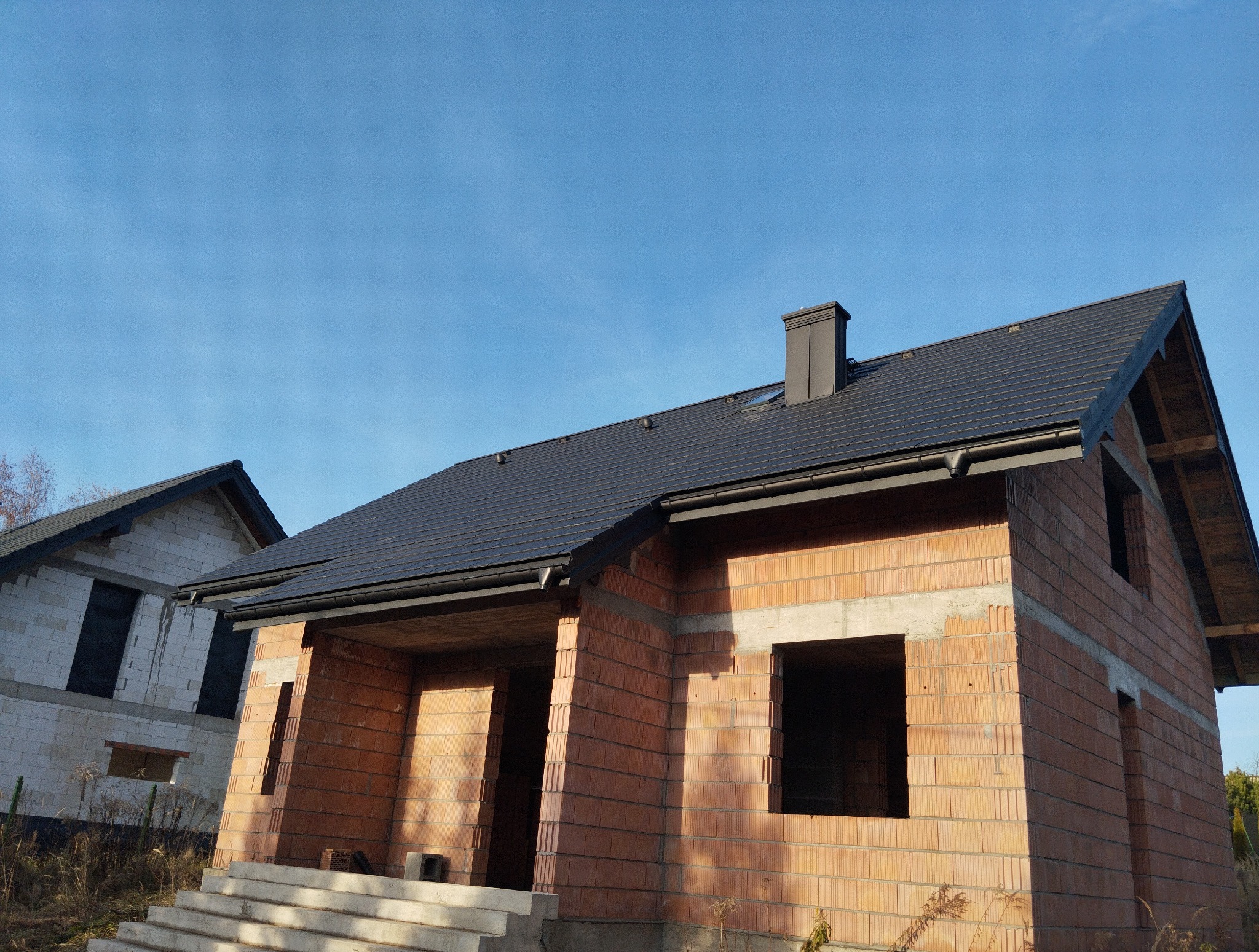 Surowy stan domu z czerwonej cegły z czarnym dachem i kominem w słoneczny dzień. Widoczne okna i drzwi bez ram. Drugi budynek w tle.