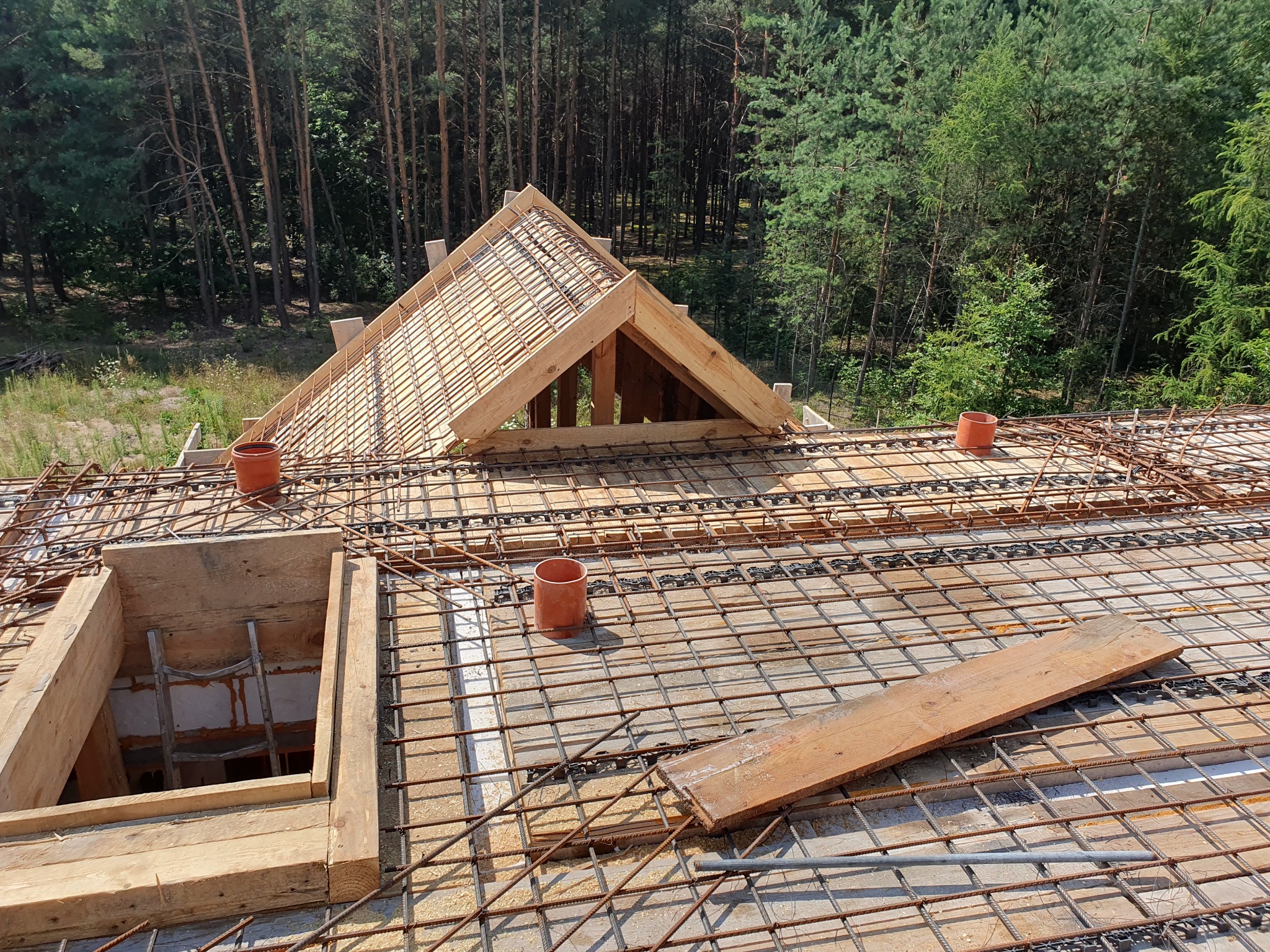 Zbrojenie stropu z drewnianą konstrukcją więźby dachowej na tle lasu, widok z góry. Prace budowlane w toku, zbrojenie z siatki stalowej, drewniane szalunki.