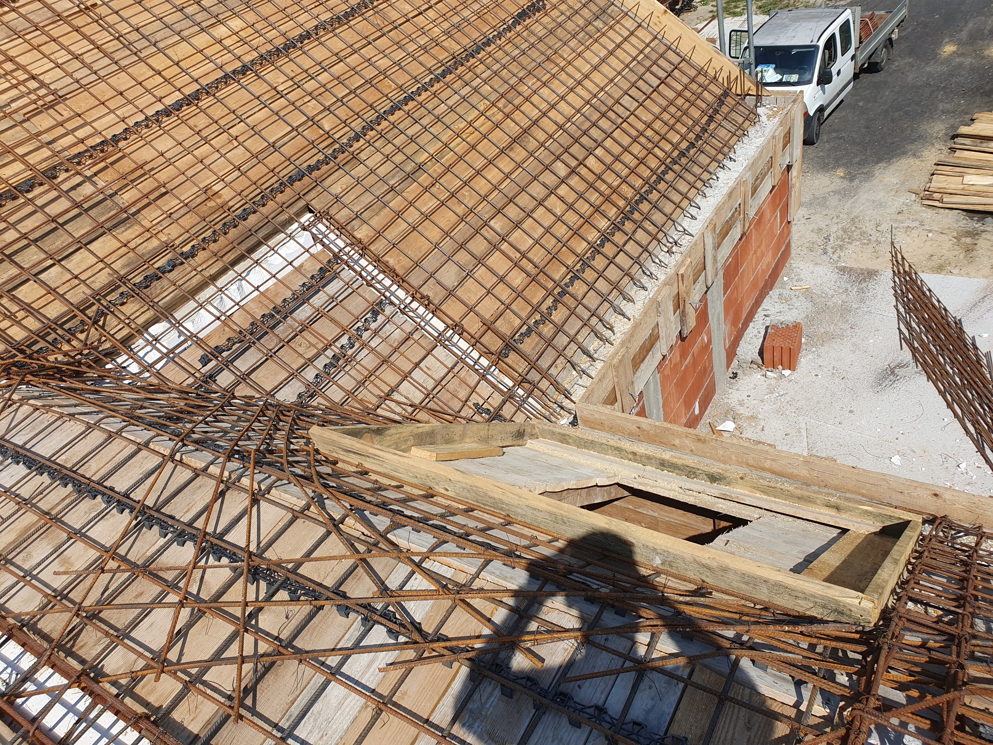 Zbrojenie stropu na drewnianym szalunku, widok z góry. Na drugim planie ściana z czerwonej cegły i biały samochód dostawczy. Cień osoby na pierwszym planie.