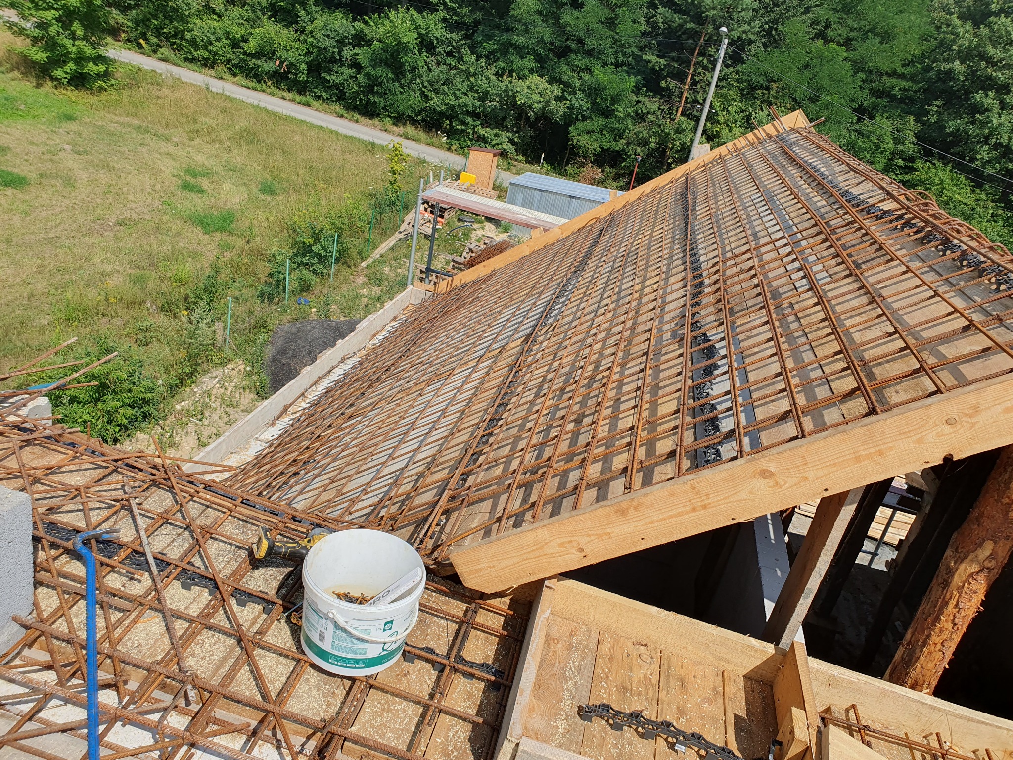 Zbrojenie dachu domu w trakcie budowy, widok z góry na siatkę z prętów stalowych, drewniane elementy konstrukcyjne i wiadro z narzędziami. Tło: zieleń drzew.