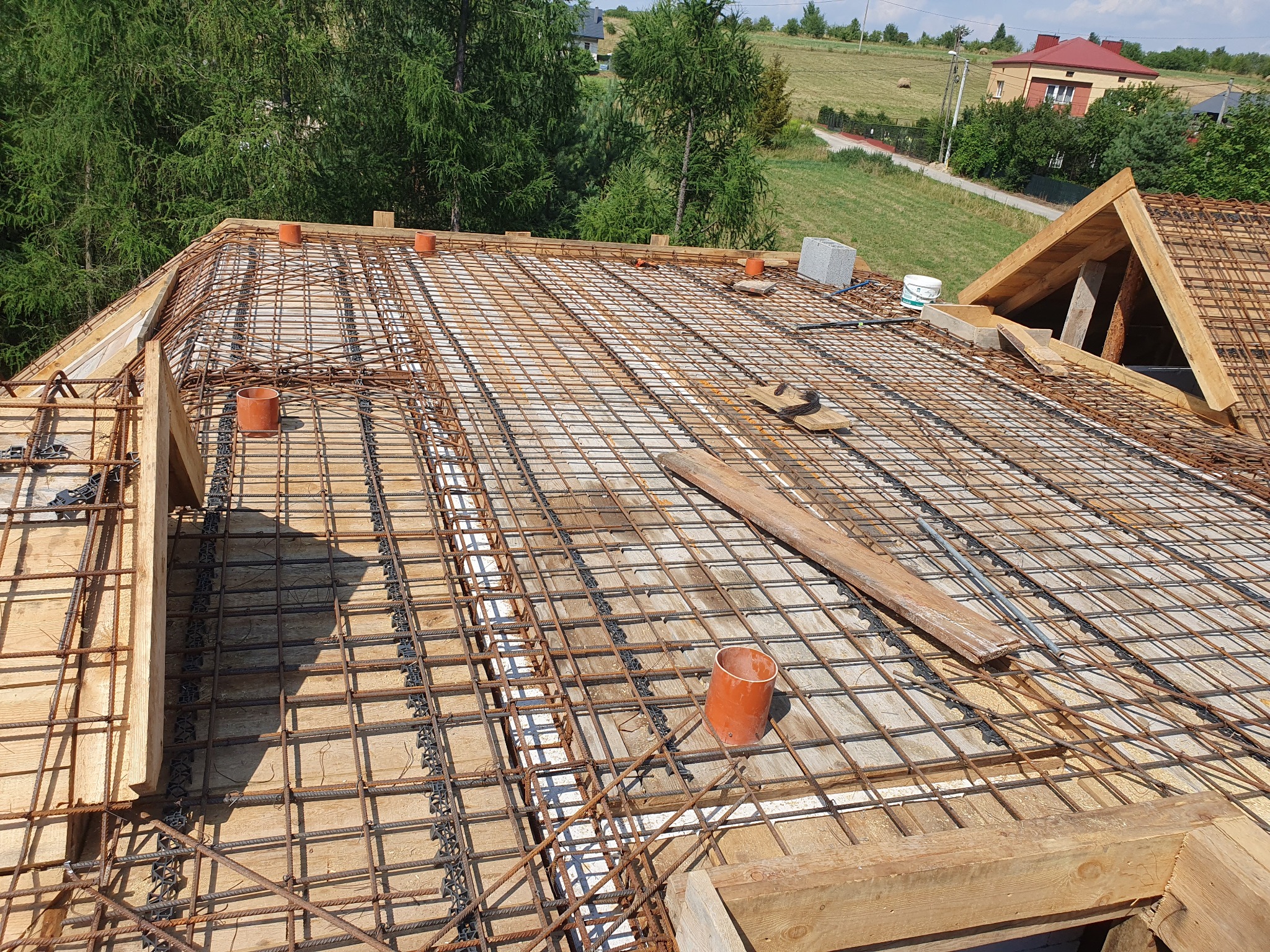 Zbrojenie dachu z siatki stalowej, drewniane szalunki, rury wentylacyjne. Widok z góry na konstrukcję przygotowaną do zalania betonem, w tle zieleń i zabudowa.