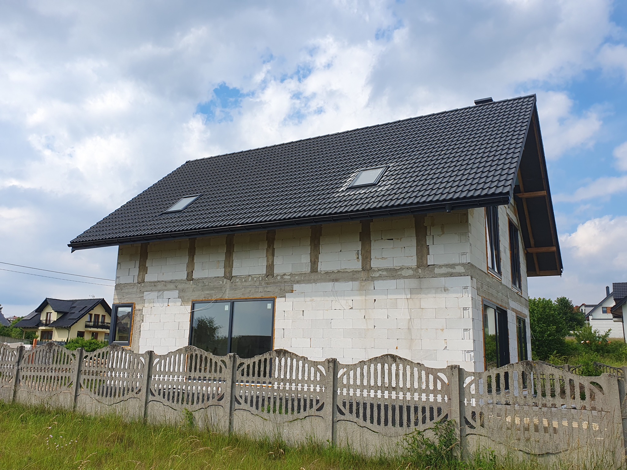 Dom w trakcie budowy z białych bloczków, czarny dach z oknami połaciowymi, betonowe ogrodzenie, trawa. Widok z zewnątrz na budynek w stanie surowym.